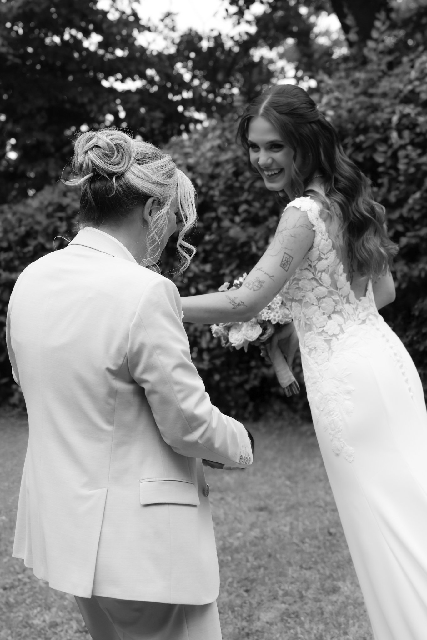 Black and white photo of two women at a wedding, holding hands and smiling at each other outdoors, with trees in the background.