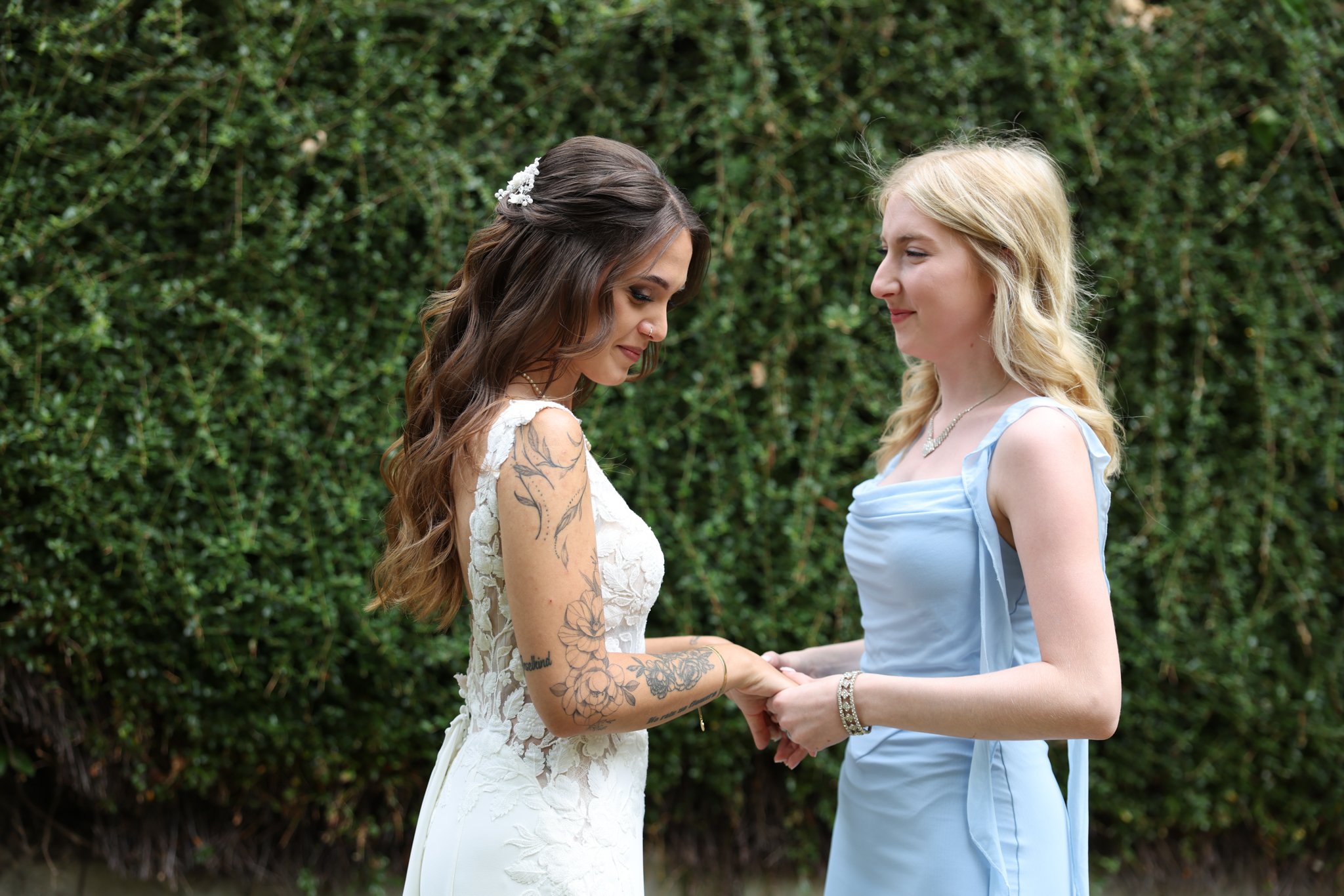 Two women, one with long brown hair in a wedding dress and the other with blonde hair in a light blue dress, holding hands outdoors in front of green bushes.