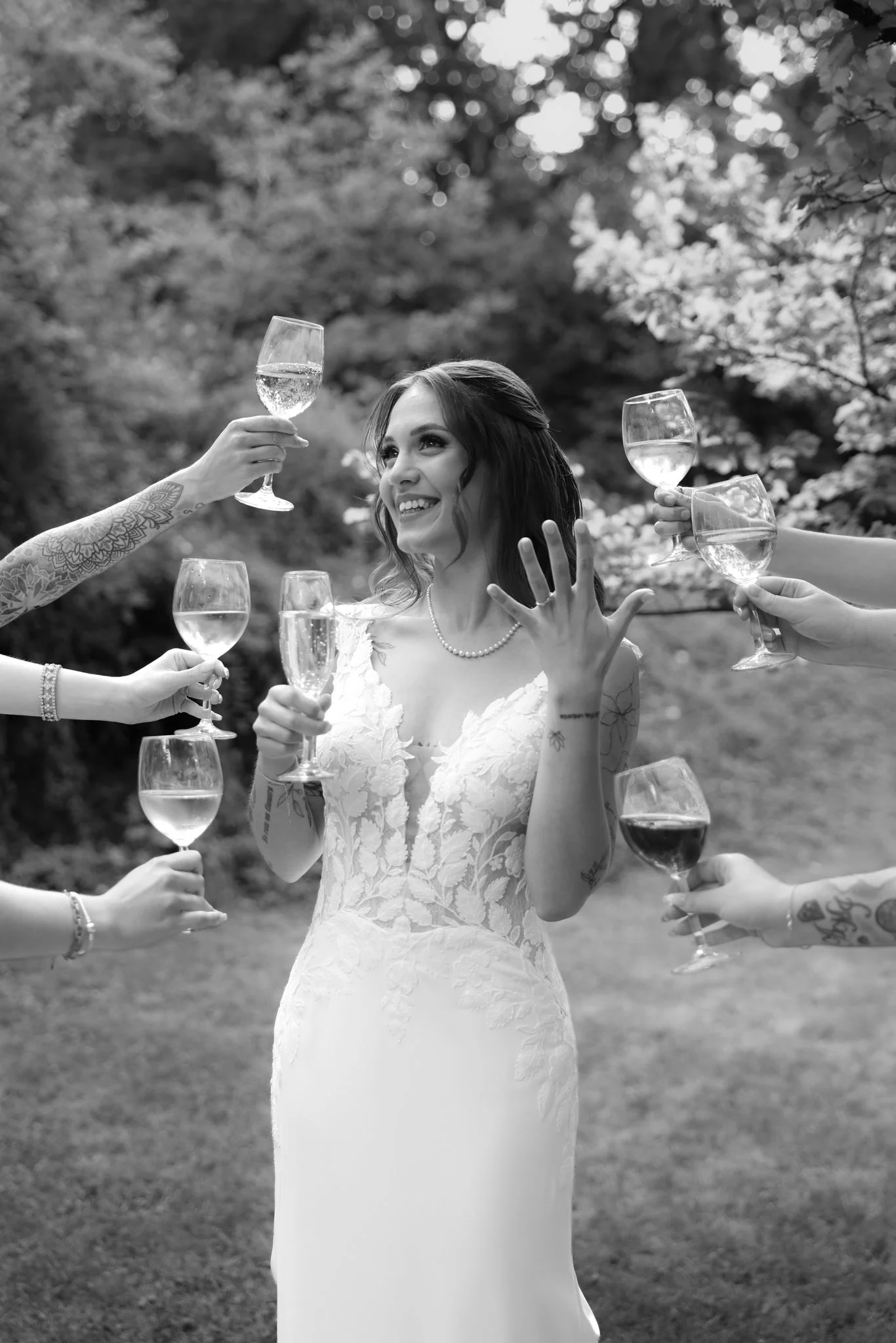A woman in a wedding dress laughing and raising her hand with a wedding band while surrounded by hands holding glasses of champagne for a toast outdoors.