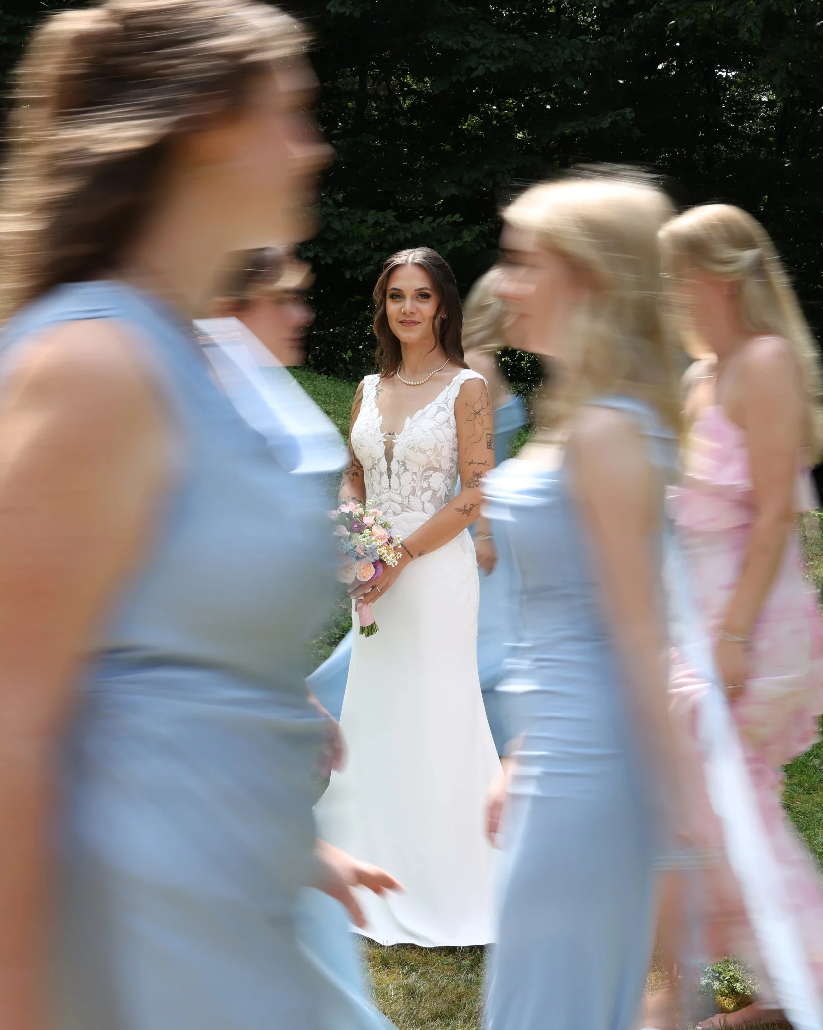 A bride in a white lace wedding dress holding a bouquet of pink and white flowers stands outdoors. Several women in pastel dresses walk past in a blur, creating motion around her. Background features green trees and grass.