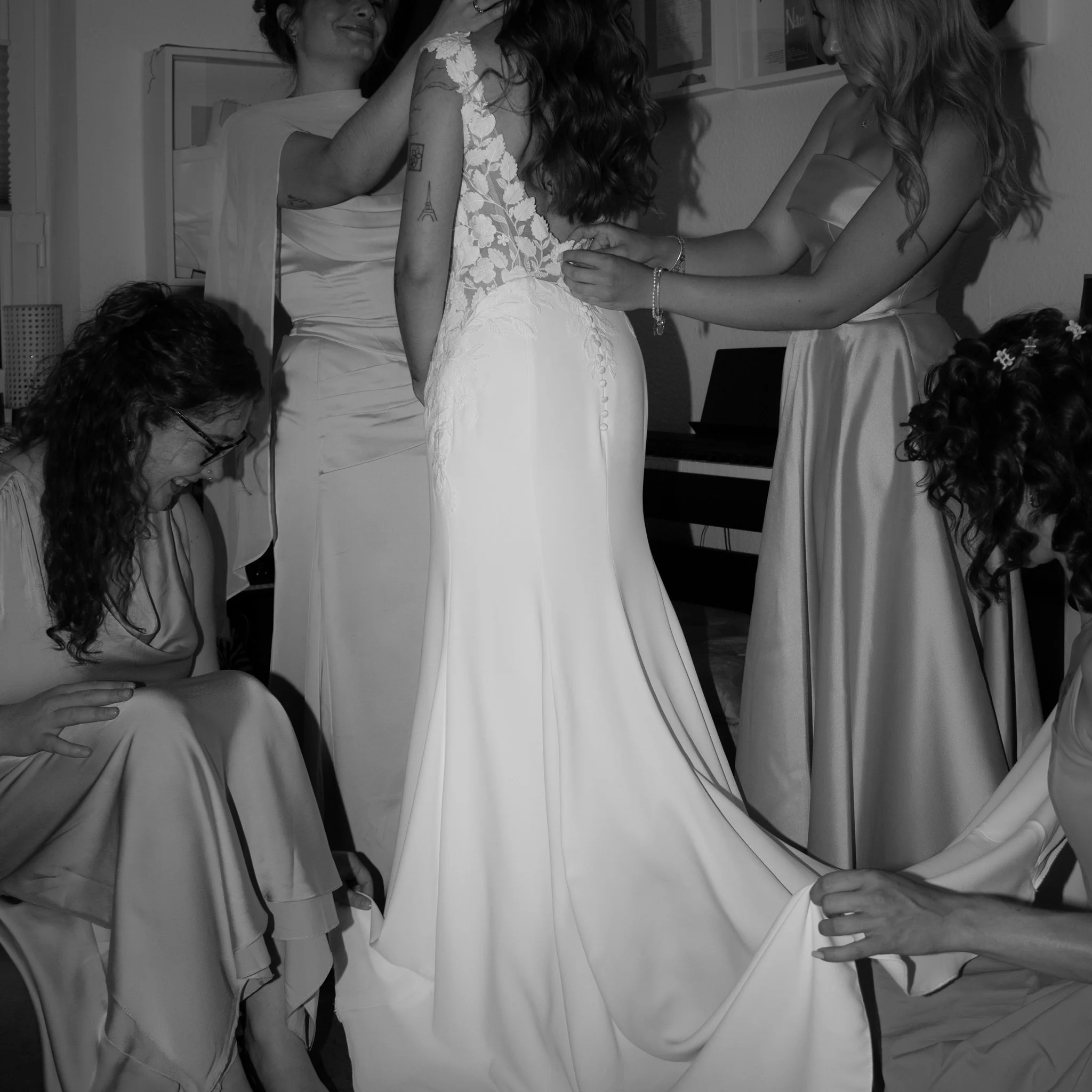 Braut wird von ihren Brautjungfern beim Anziehen des Hochzeitskleides unterstützt.