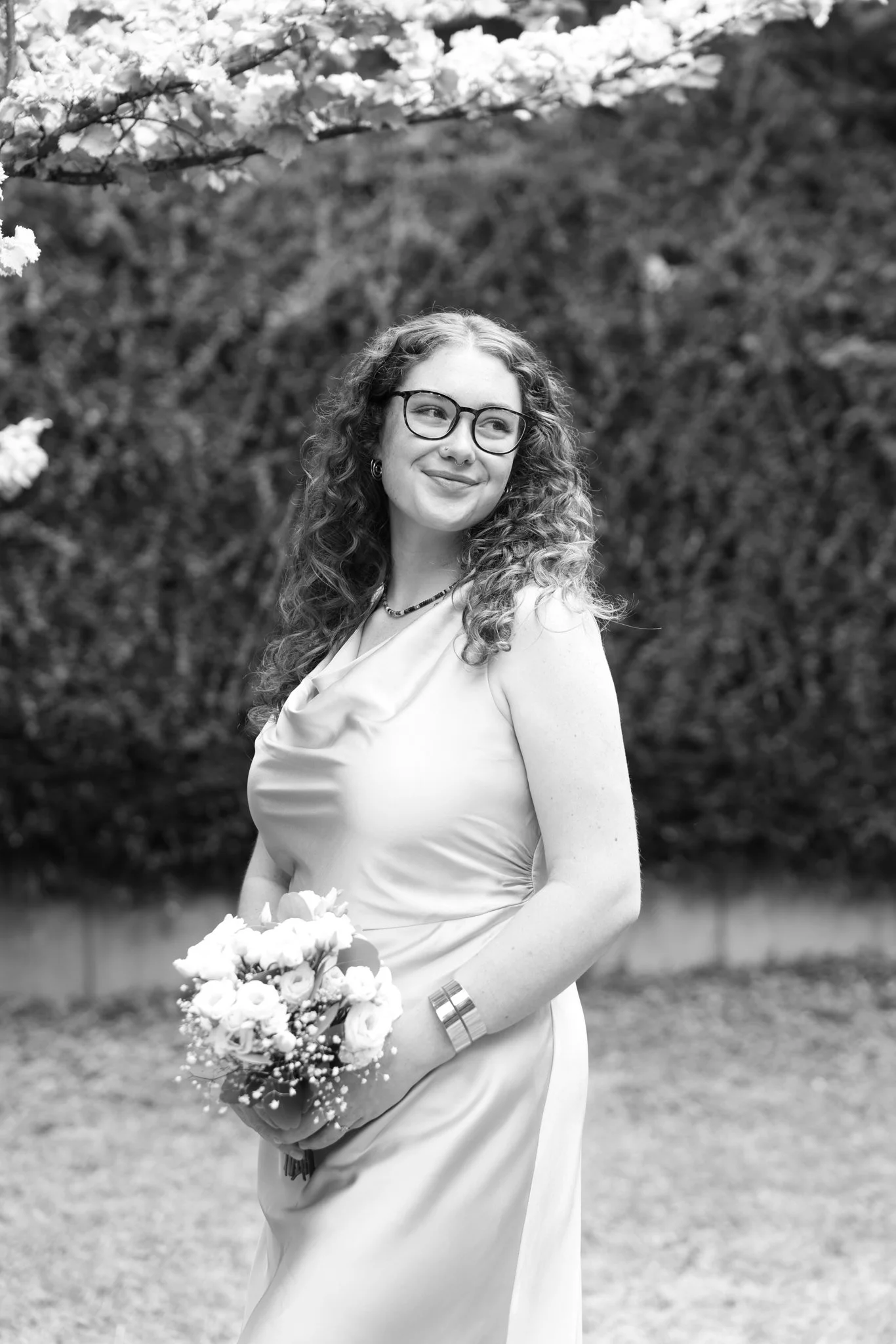 Brautjungfer mit lockigen Haaren und Brille hält einen Blumenstrauß im Freien.