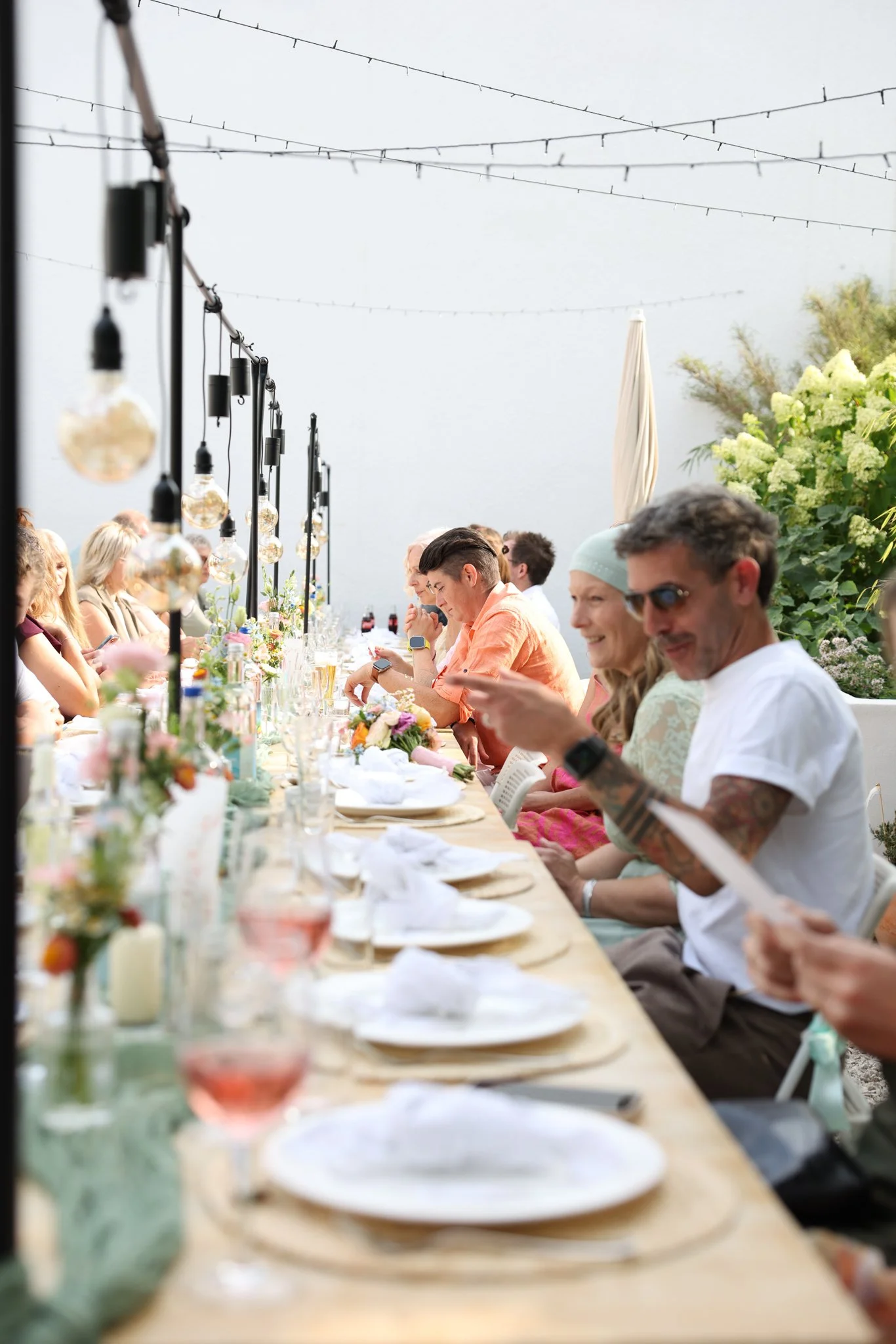 People sitting at a long outdoor dining table decorated with flowers, with string lights hanging overhead and greenery in the background.