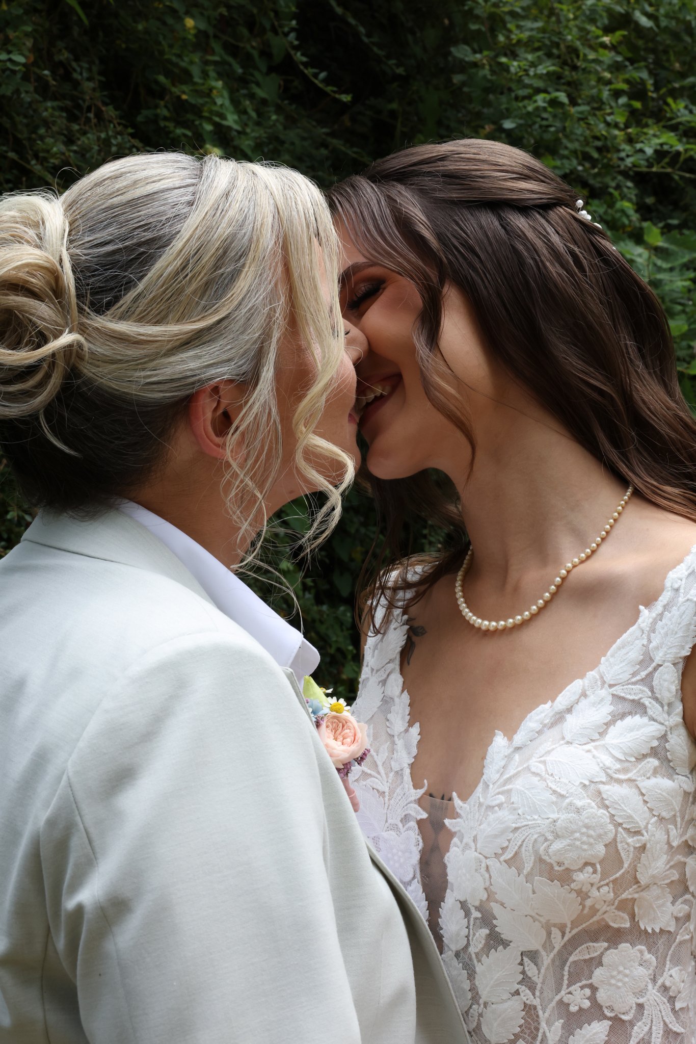 Two women dressed in wedding attire, touching foreheads and smiling, standing outdoors with green foliage in the background.