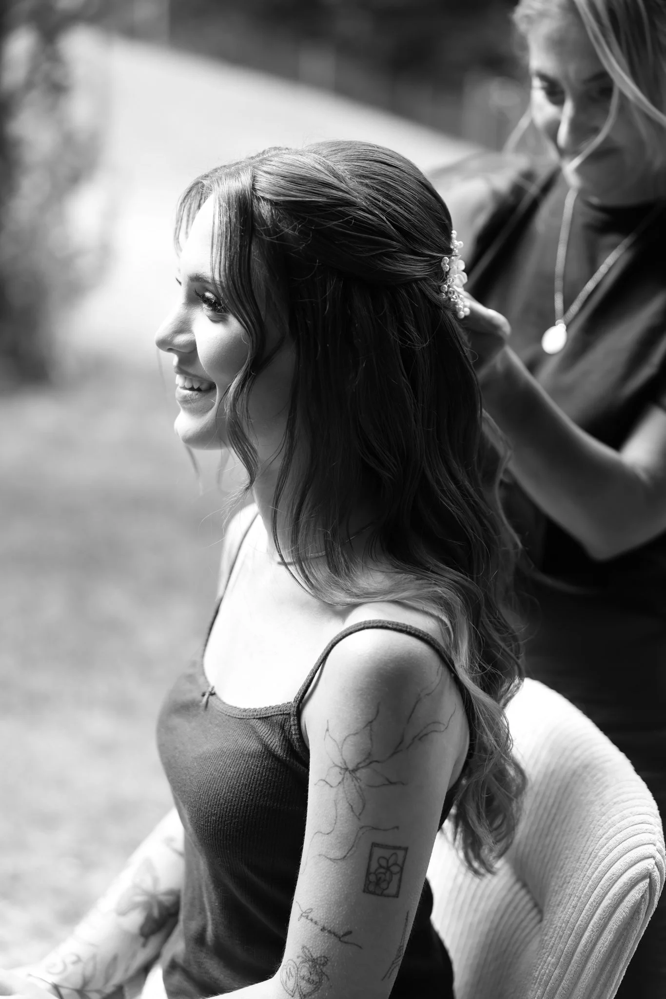 A woman with long wavy hair smiling, sitting outdoors with another woman helping her with her hair, which is adorned with a decorative hairpin. The woman has tattoos on her arm, including a flower and a garden scene, and is wearing a tank top.