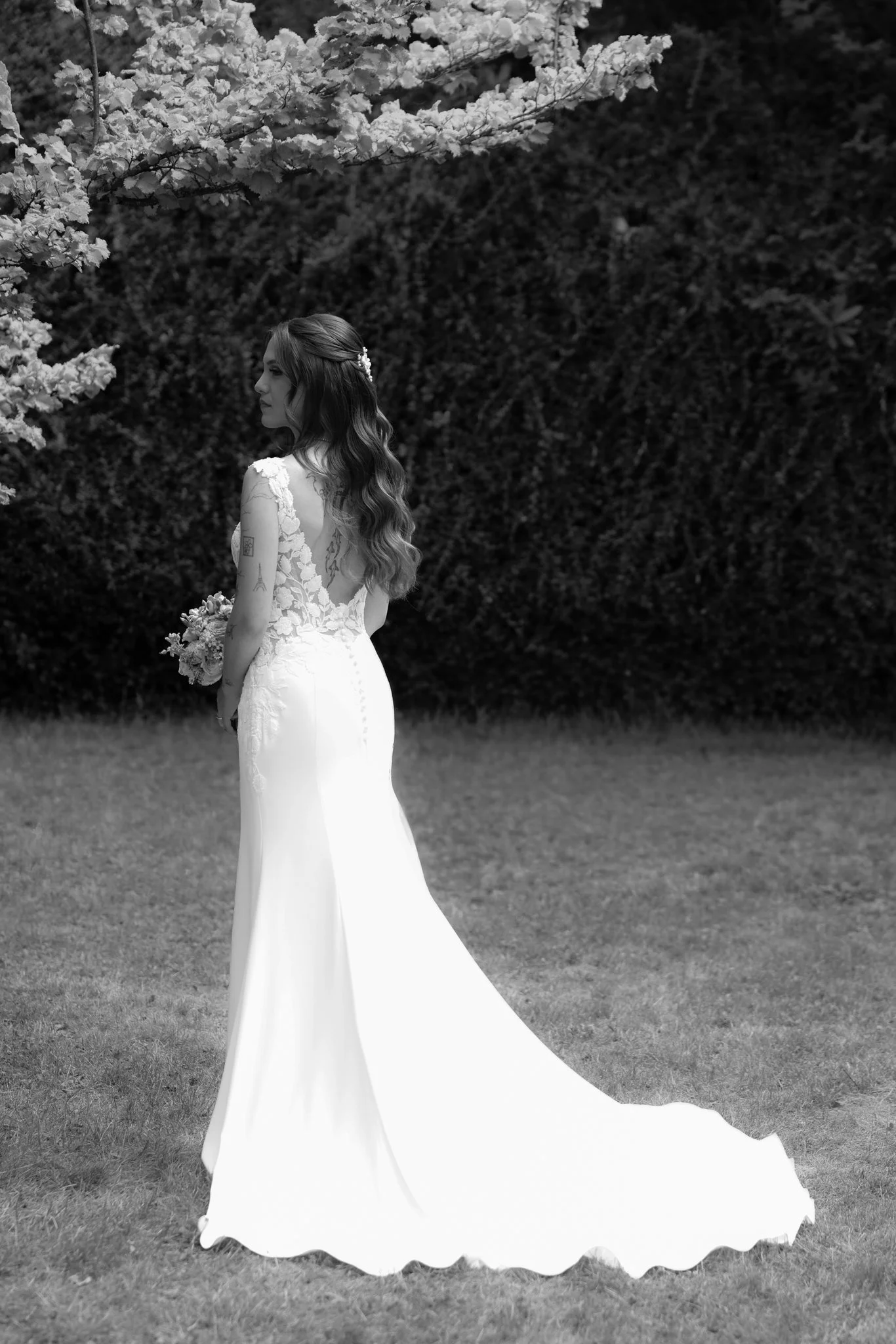 A woman in a wedding dress holding a bouquet, standing outdoors on grass with a hedge in the background and a flowering tree overhead.