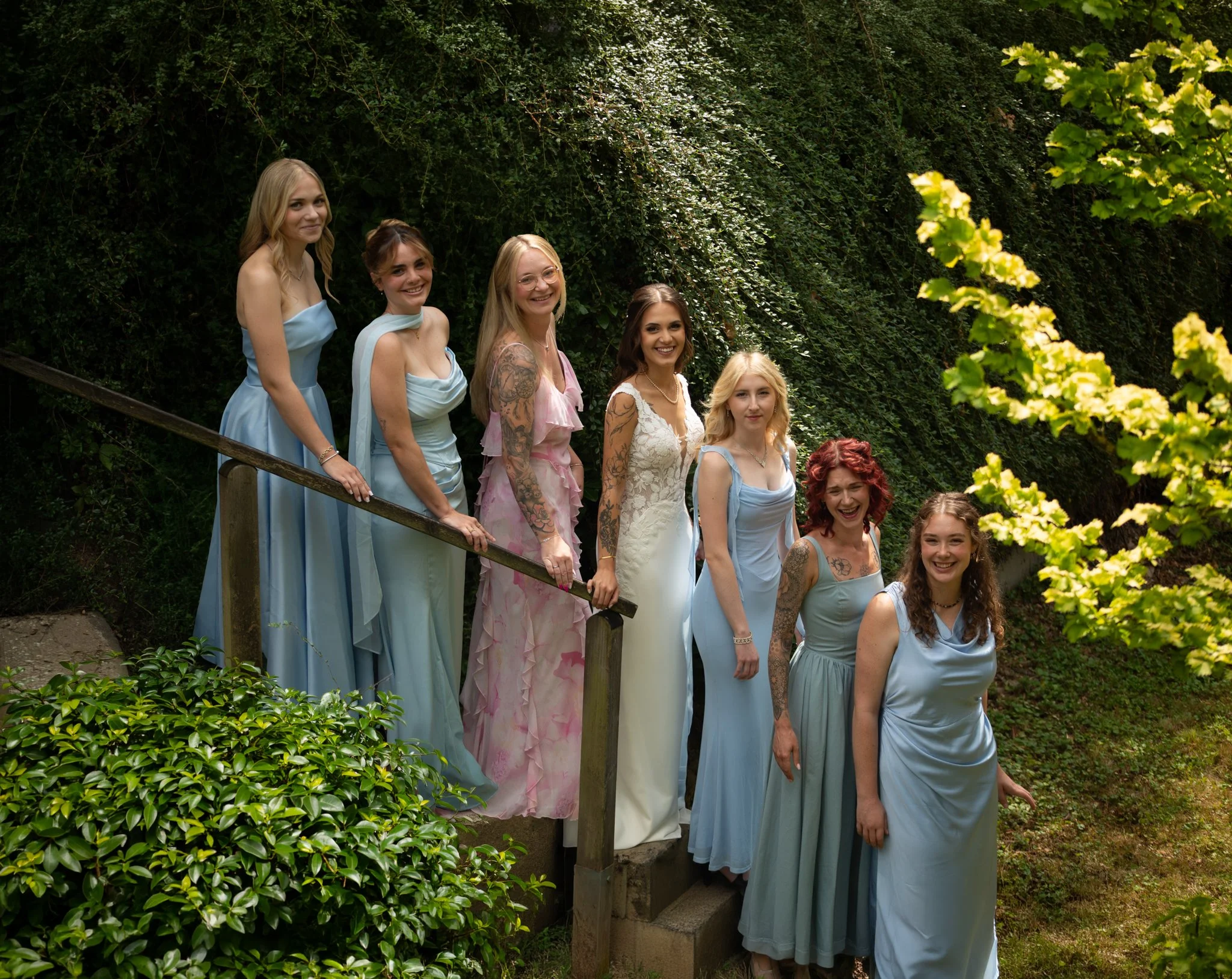 Braut und sechs Brautjungfern auf einer Holztreppe im Grünen – Gruppenportrait vor der Trauung.