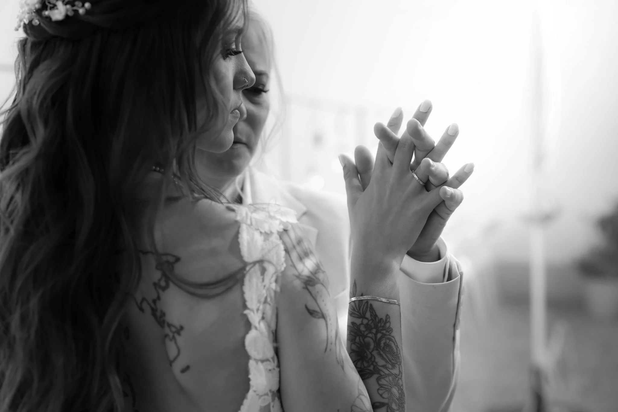 A woman with long dark hair and tattoos on her arm and hand, wearing a ring and a bracelet, is looking at her fingers while posing with her hands in front of her face.