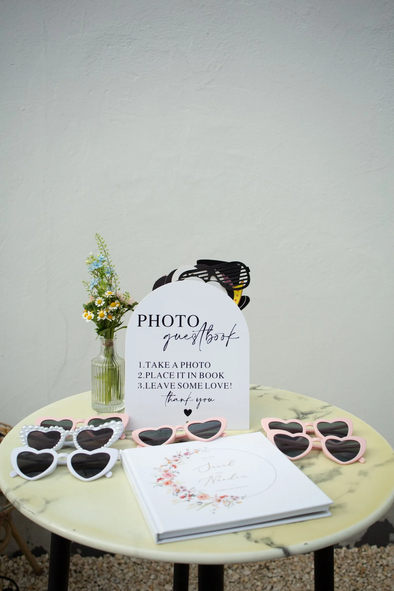 A table with various decorative sunglasses, a white guestbook with floral design, a sign with instructions for guests, a small vase of flowers, and a guest book with the name Sarah Nolan in cursive.