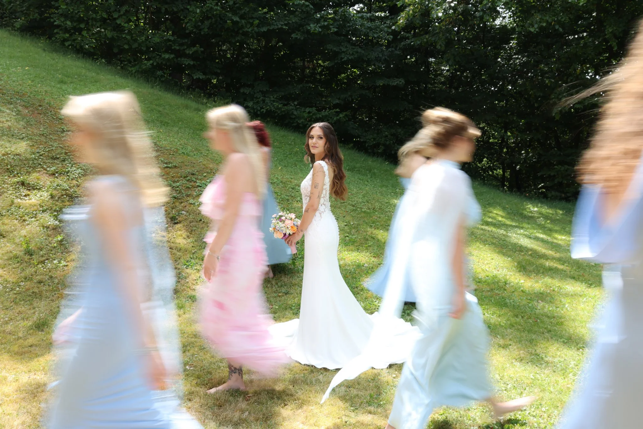 A bride standing on grass holding a bouquet in front of a blurred procession of women in colorful dresses, with trees in the background.