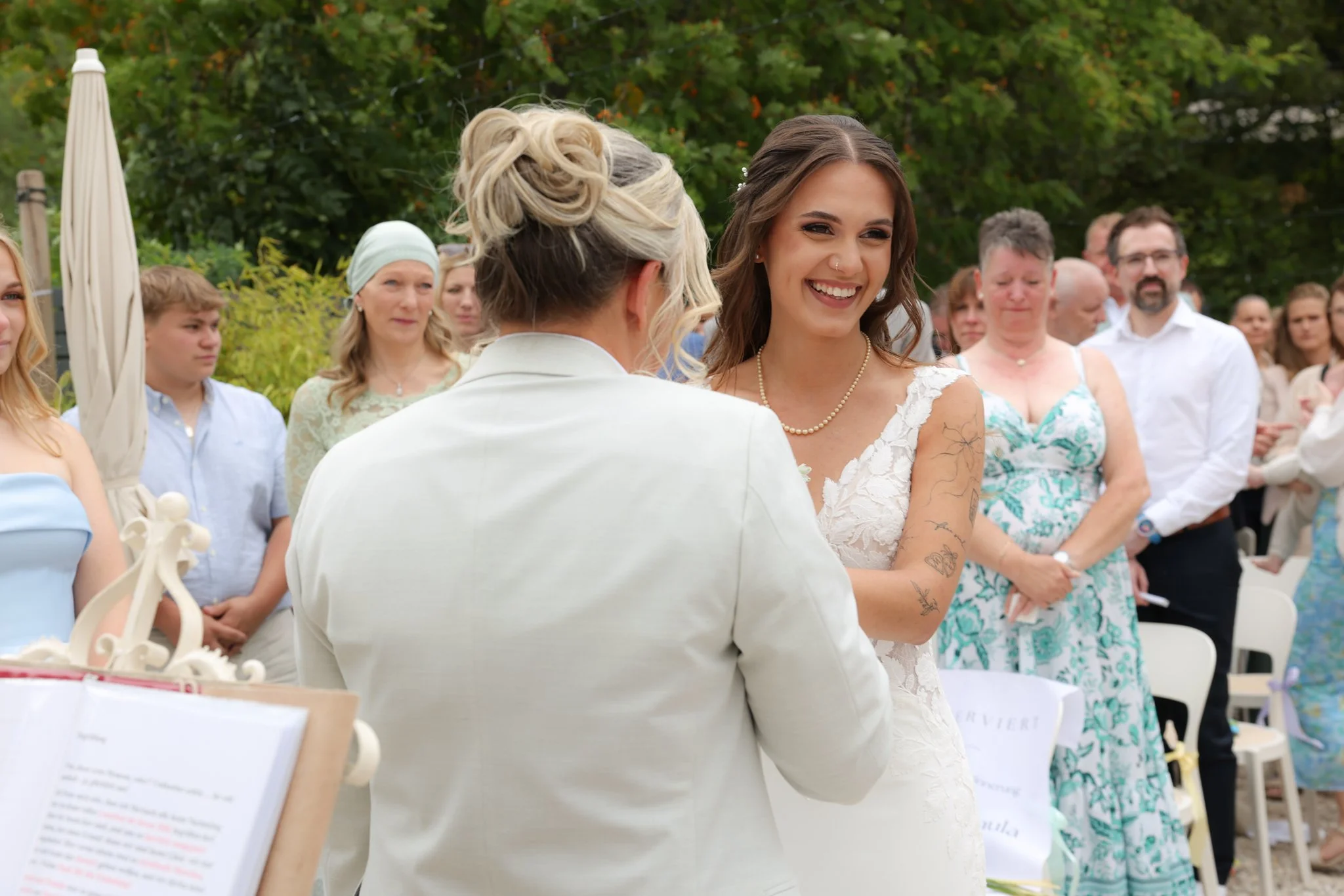 A bride with tattoos on her arms smiling at a woman during a wedding ceremony outdoors surrounded by guests.