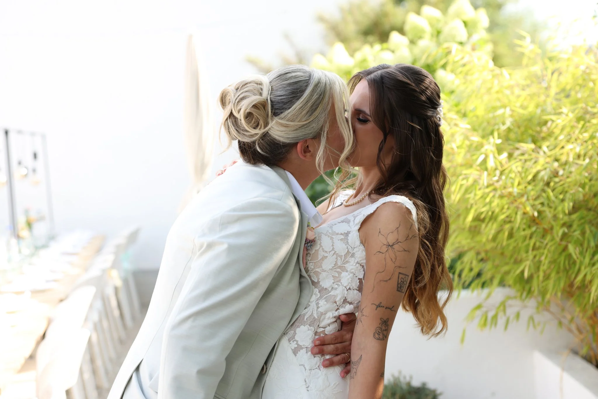 Two women, one with blonde hair and the other with dark brown hair, share a kiss outdoors, dressed in wedding attire, with greenery and white chairs in the background.