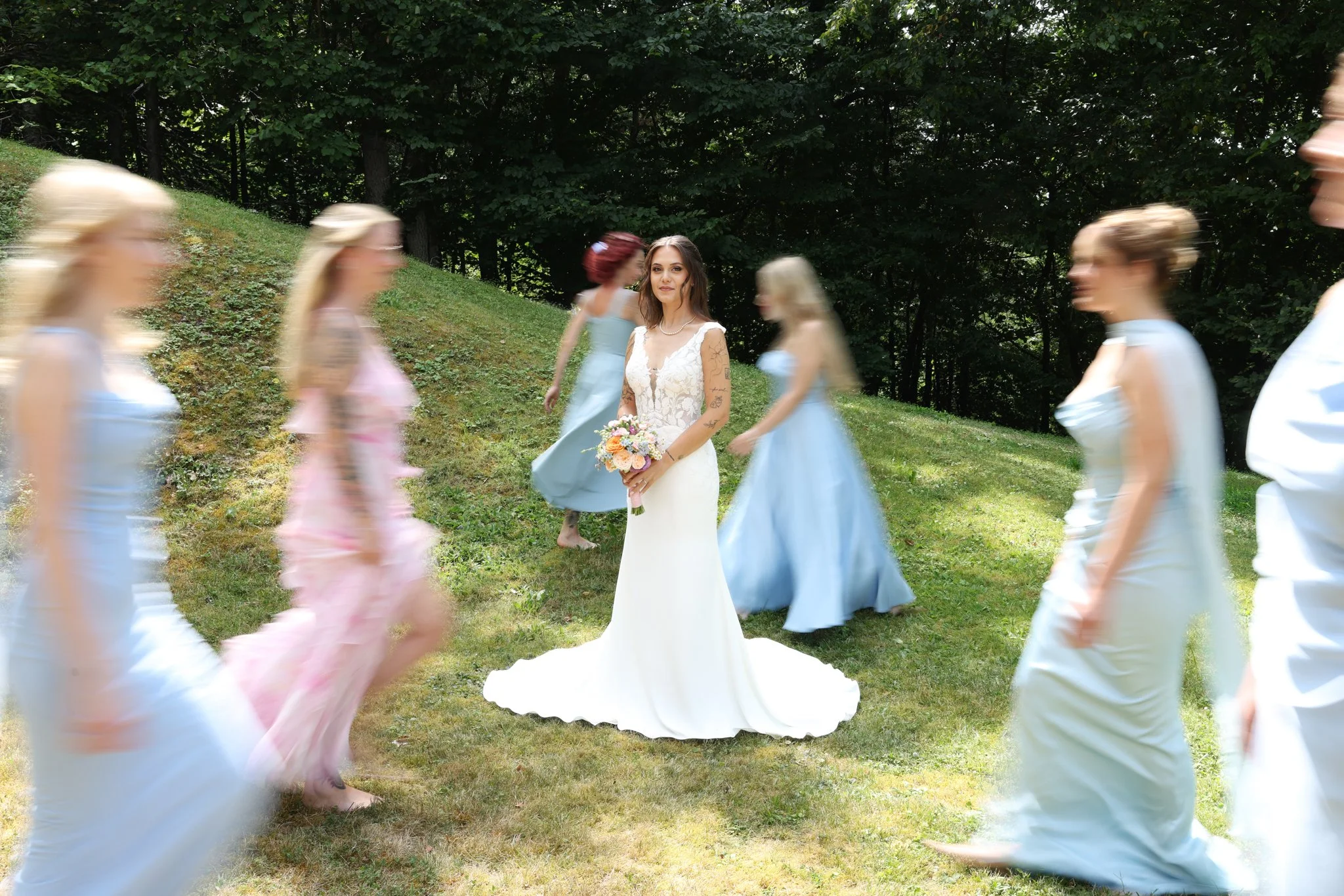 A woman in a white wedding dress holding a bouquet stands in the center while women in pastel-colored dresses walk around her in a circle outdoors, with trees in the background.