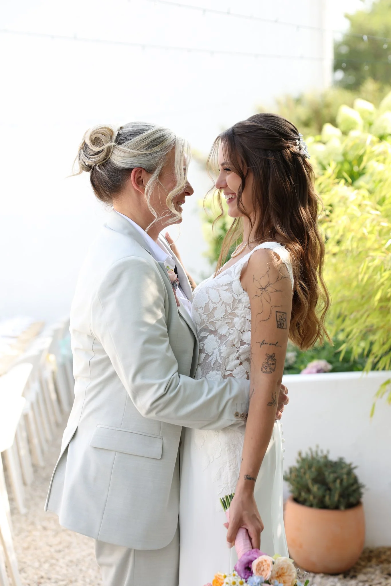 Two women, one with blonde hair in a bun dressed in a cream tuxedo and one younger with brown hair in a braid dressed in a white lace dress, face each other affectionately at an outdoor wedding or similar celebration, holding hands and smiling.