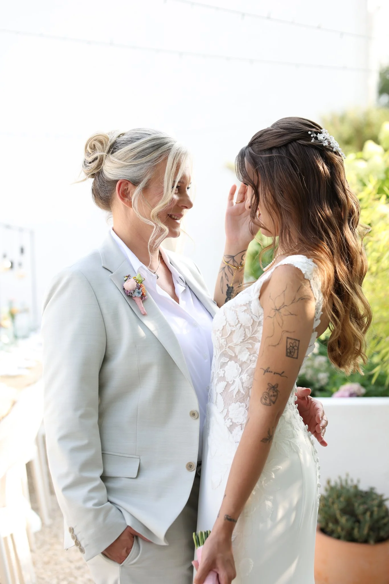 Two women, one older with blonde hair in a bun and a light-colored suit, and a  woman with long brown hair and tattoos, wearing a white wedding dress. They are standing close, smiling, and touching foreheads during a wedding ceremony outdoors.