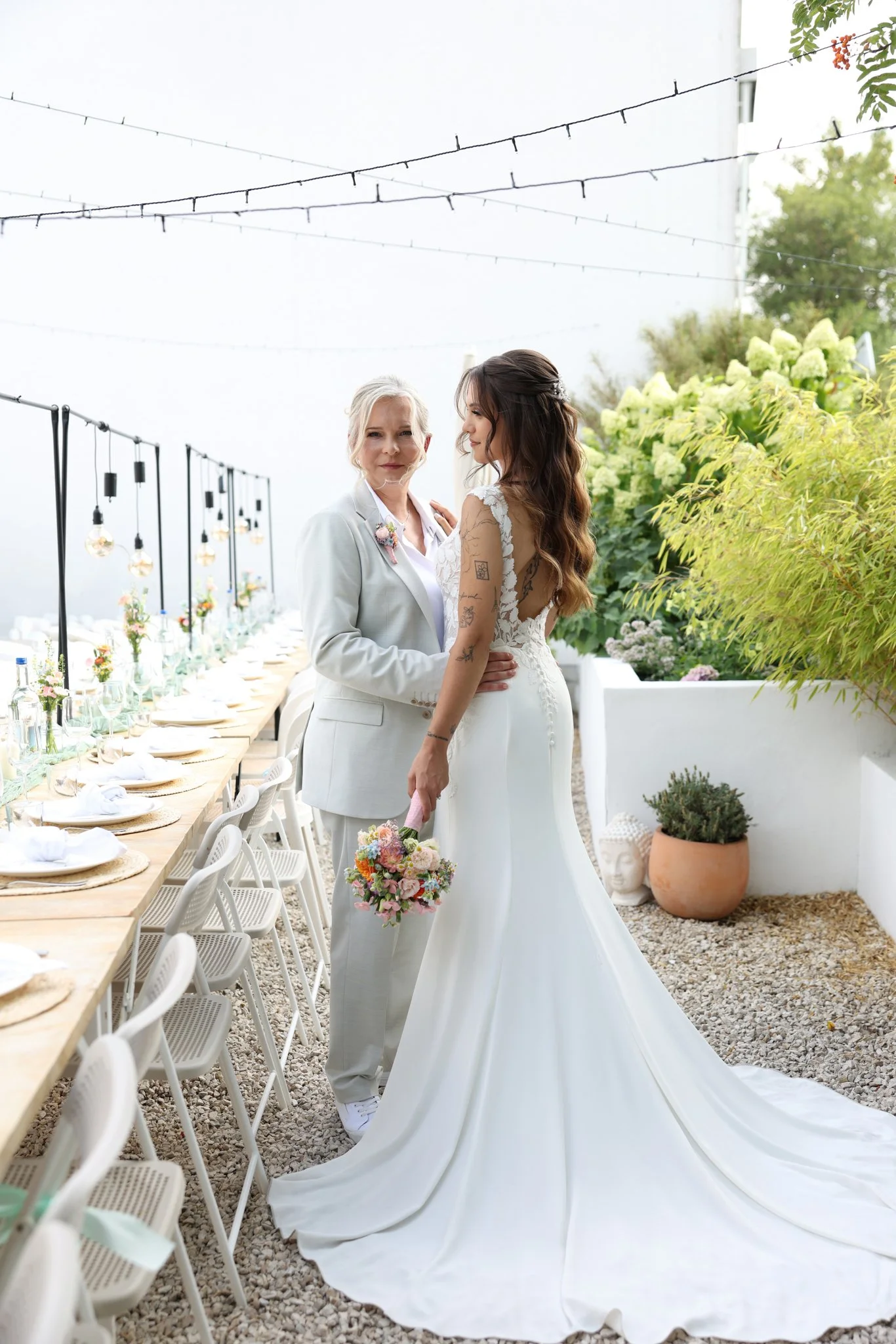 A bride in a white wedding gown and an woman in a light grey suit sharing a moment at an outdoor wedding reception with decorated tables and string lights.
