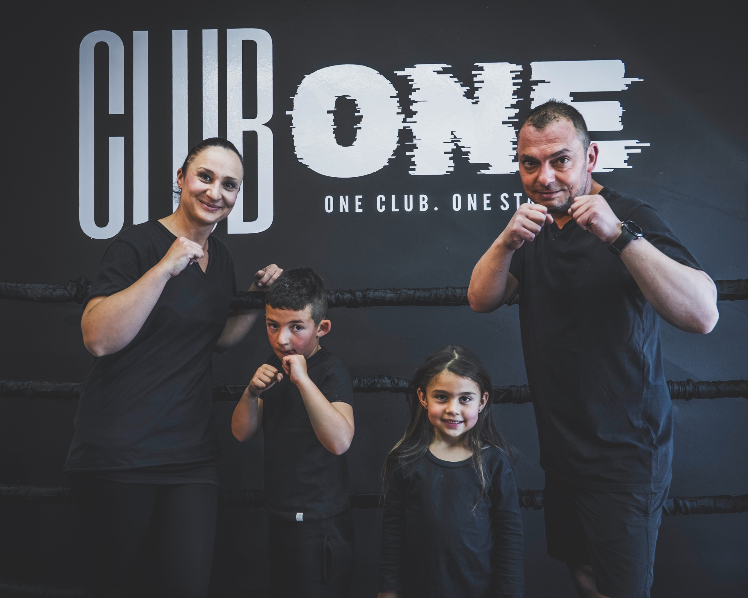 Four people, including two children, in a boxing stance inside the Club One gym with a wall sign that reads "CLUB ONE. ONE CLUB. ONE STANDARD."