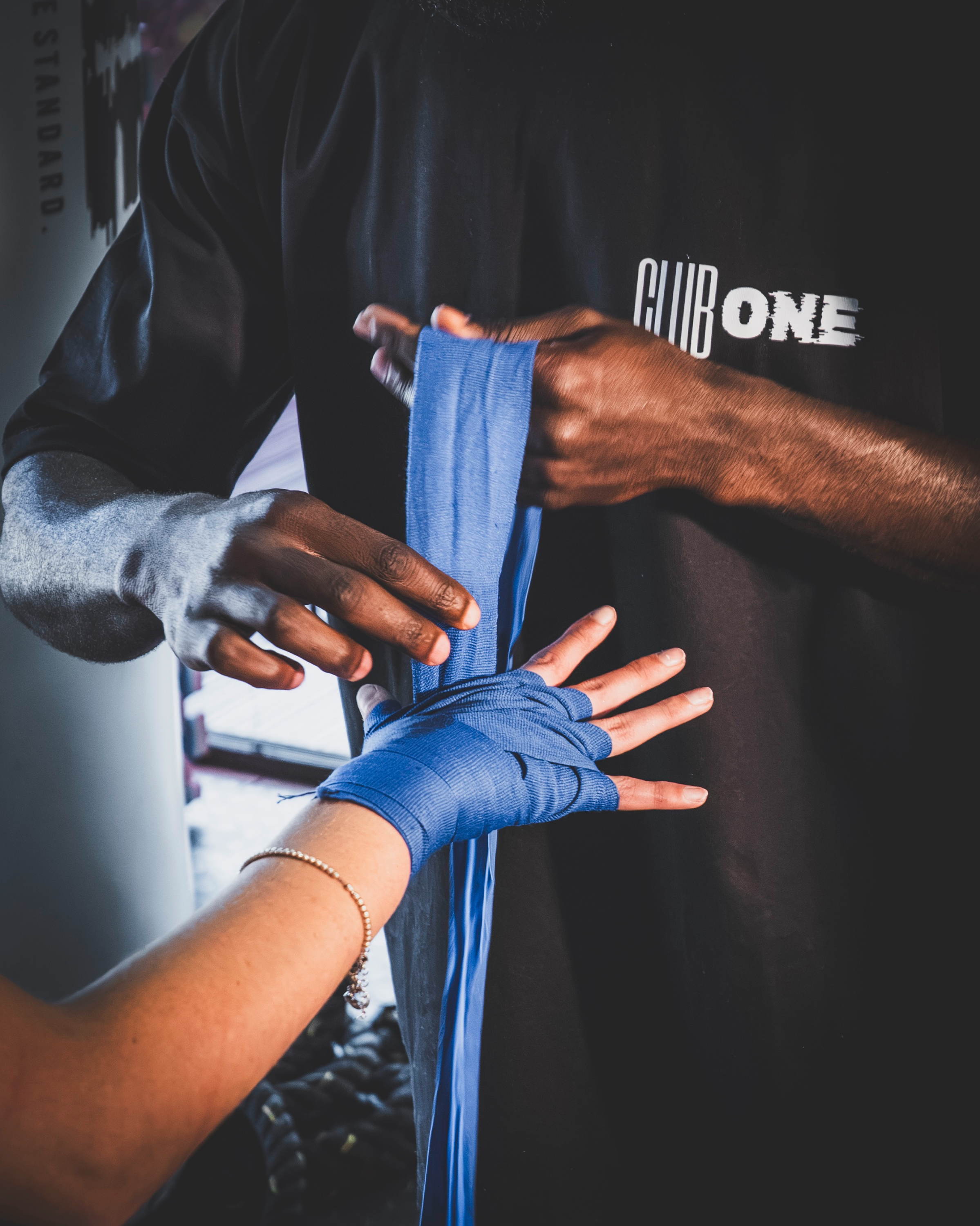 A person with long dark hair wearing a black shirt with white text 'CLUB ONE' seen from their chest up, being assisted by a person with a tan complexion, wearing a light-colored long sleeve shirt and blue hand wraps, during a boxing or martial arts class.