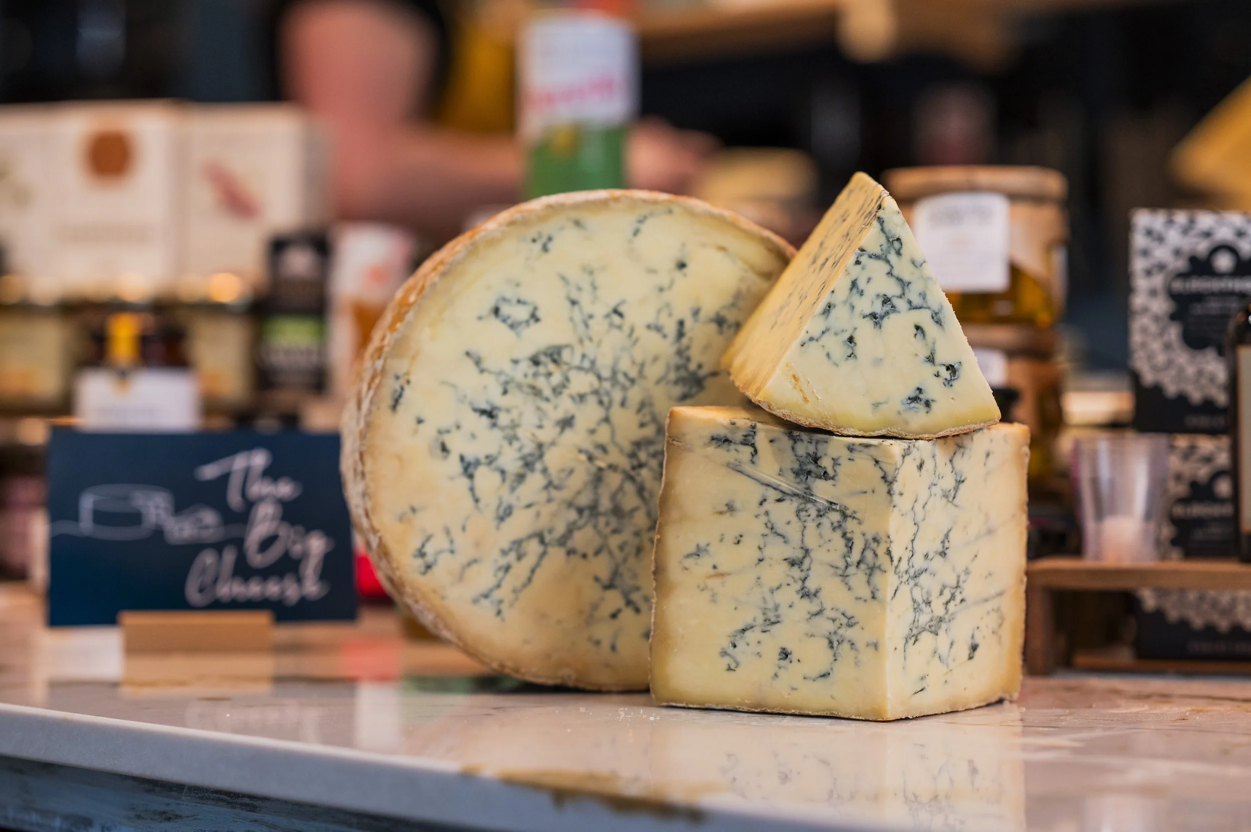 A large wheel of blue-veined cheese with a wedge cut out, displaying a marbled interior, placed on a white surface with various cheese products blurred in the background.