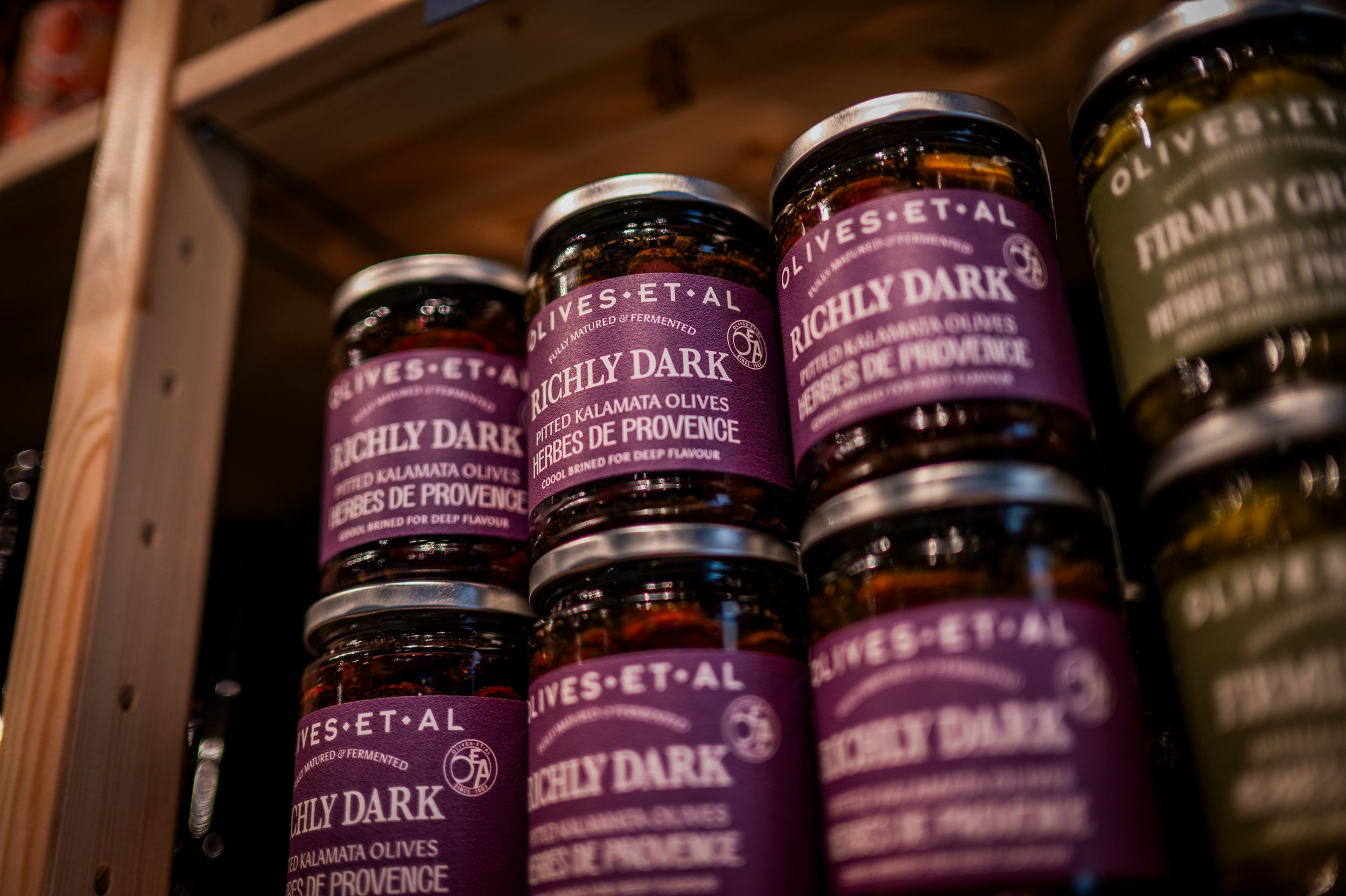 Multiple jars of olives with purple and olive green labels on a wooden shelf.