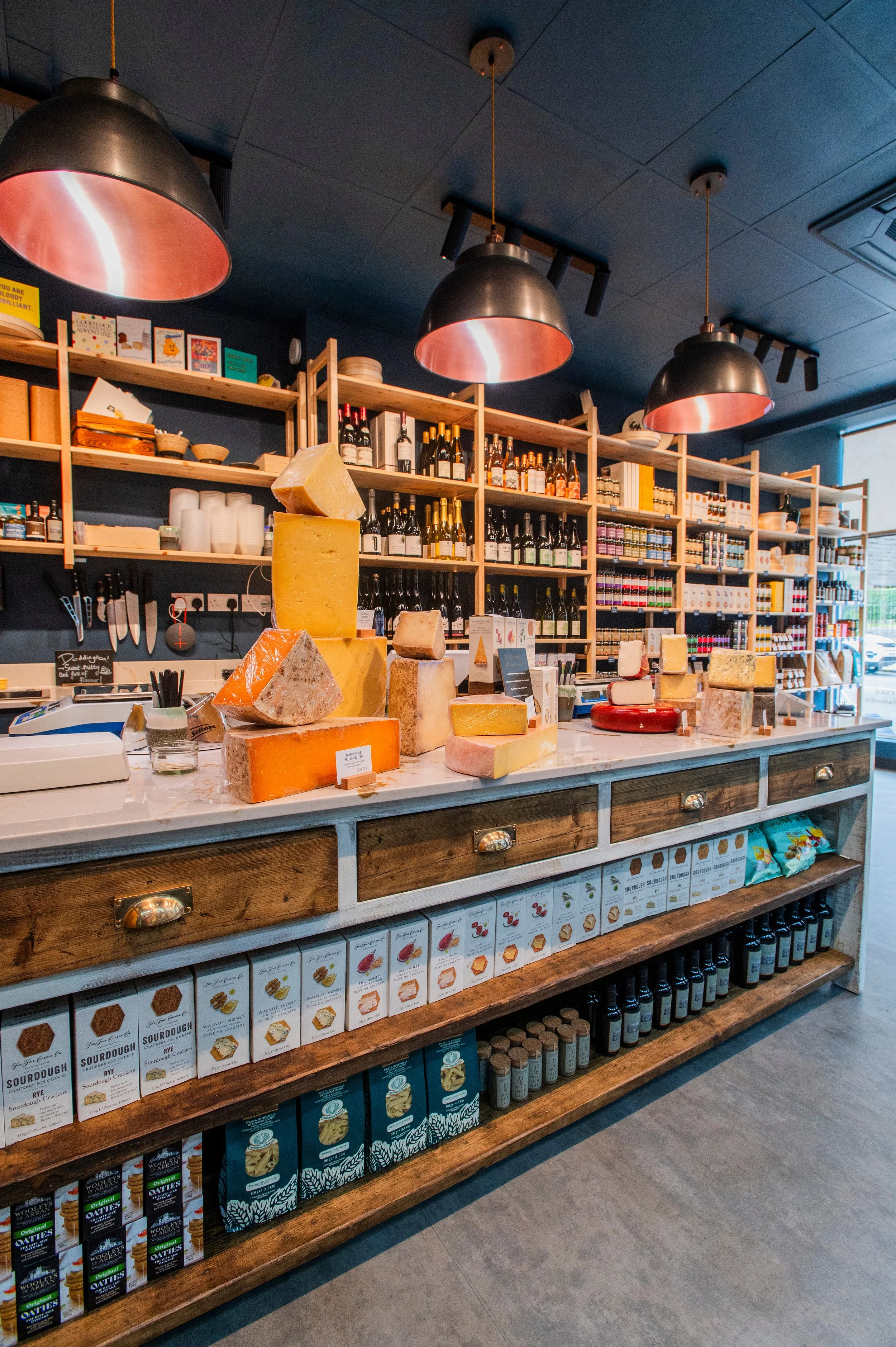 Inside a store with wooden shelves stocked with bottles, boxes, and cheeses. Several large black pendant lights hang from a dark ceiling. The countertop displays various cheeses of different sizes and styles.