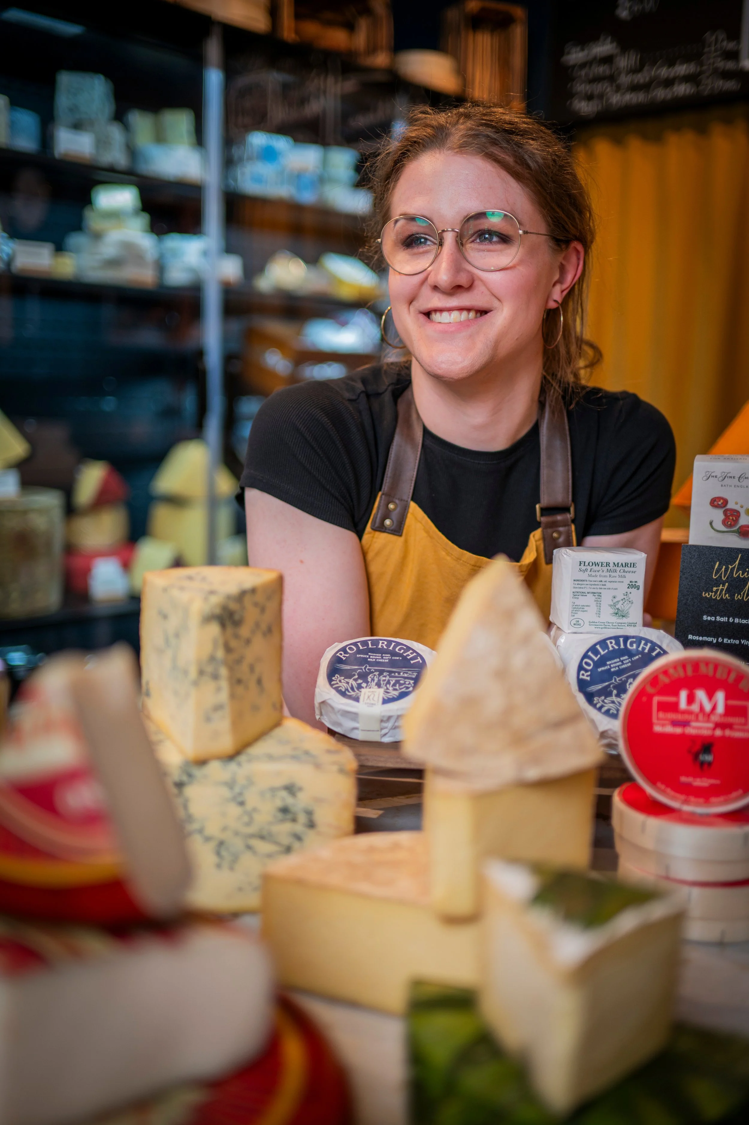 A smiling woman wearing glasses and a black shirt with a yellow apron, standing behind a cheese display at a market or shop, with various cheeses and cheese packaging visible in the foreground and background.
