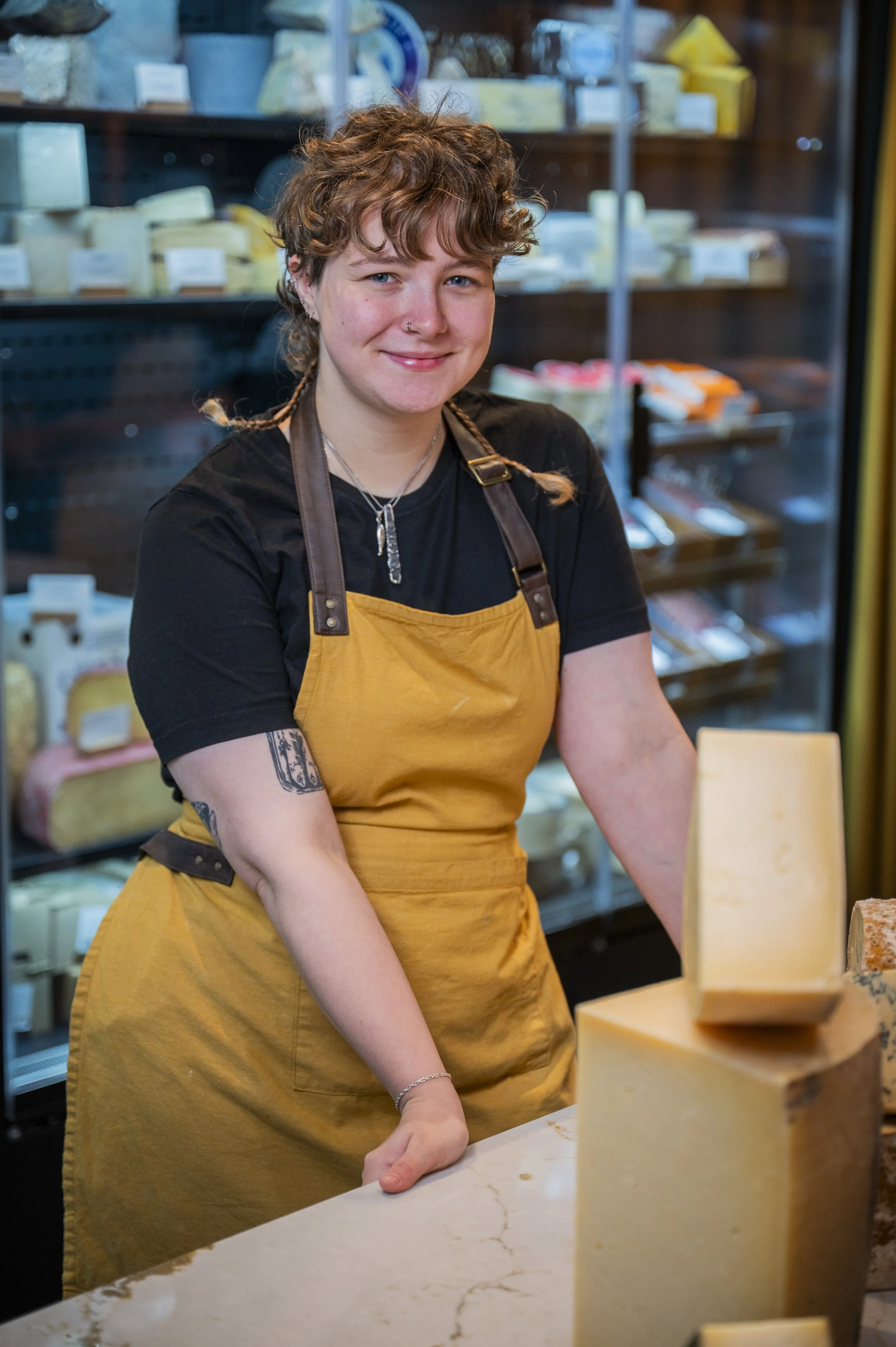 A woman with short curly hair and a nose piercing wearing a black shirt and a mustard-colored apron, standing behind a cheese display counter in a cheese shop. There are blocks of cheese on the counter and cheese products on shelves behind her.
