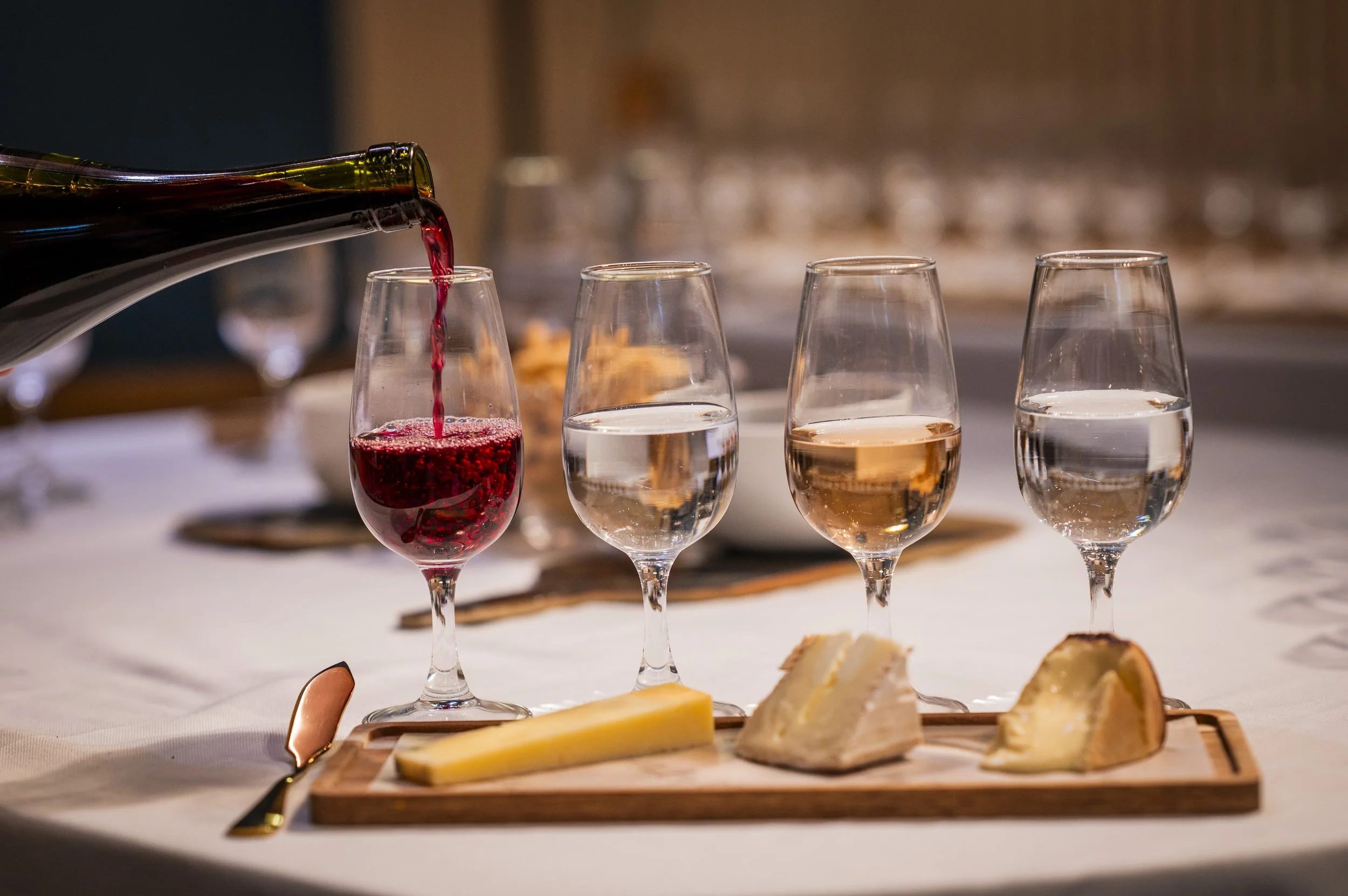 Red wine being poured into a glass with other empty glasses nearby, along with a cheese platter on a wooden board featuring various cheeses, crackers, and sliced cheese, set on a white tablecloth.