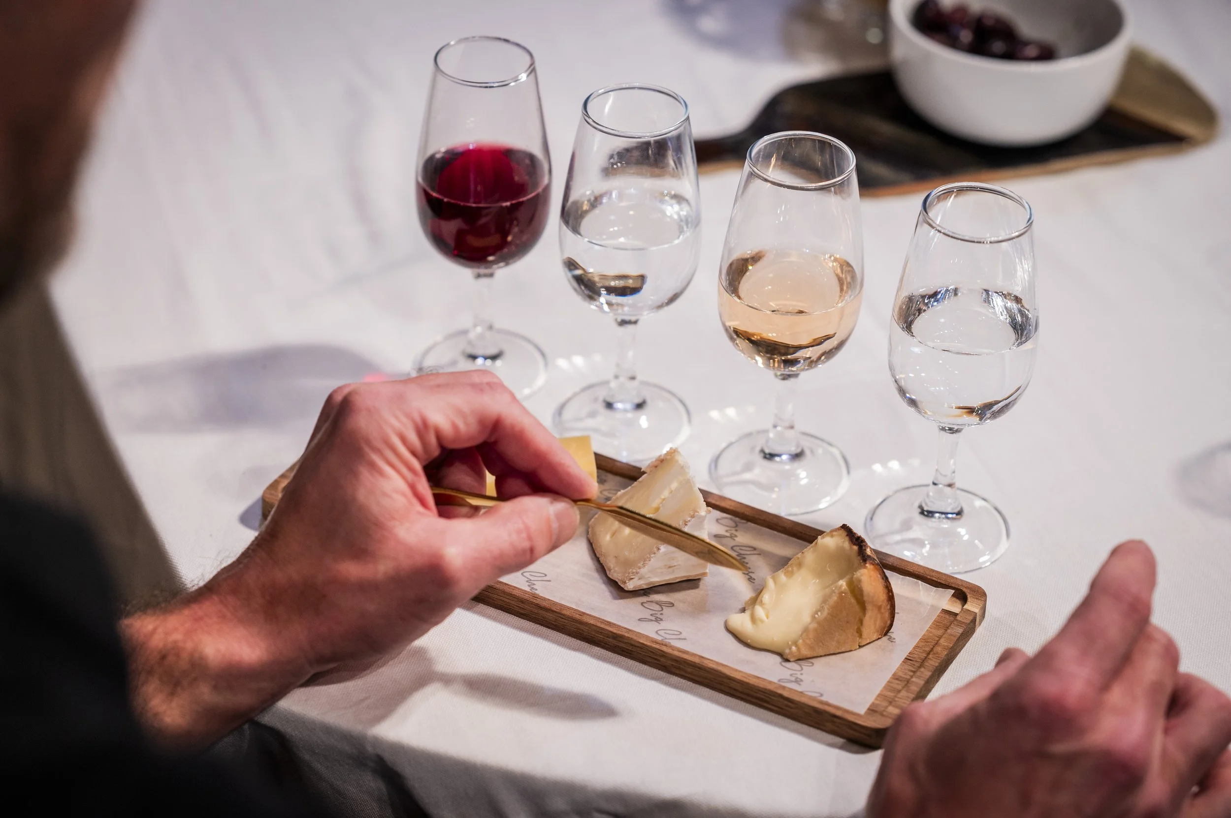 Person's hand holds a piece of cheese with a small fork, on a wooden cheese board, with four wine glasses filled with red, white, blush, and clear drinks in the background, arranged on a white tablecloth.