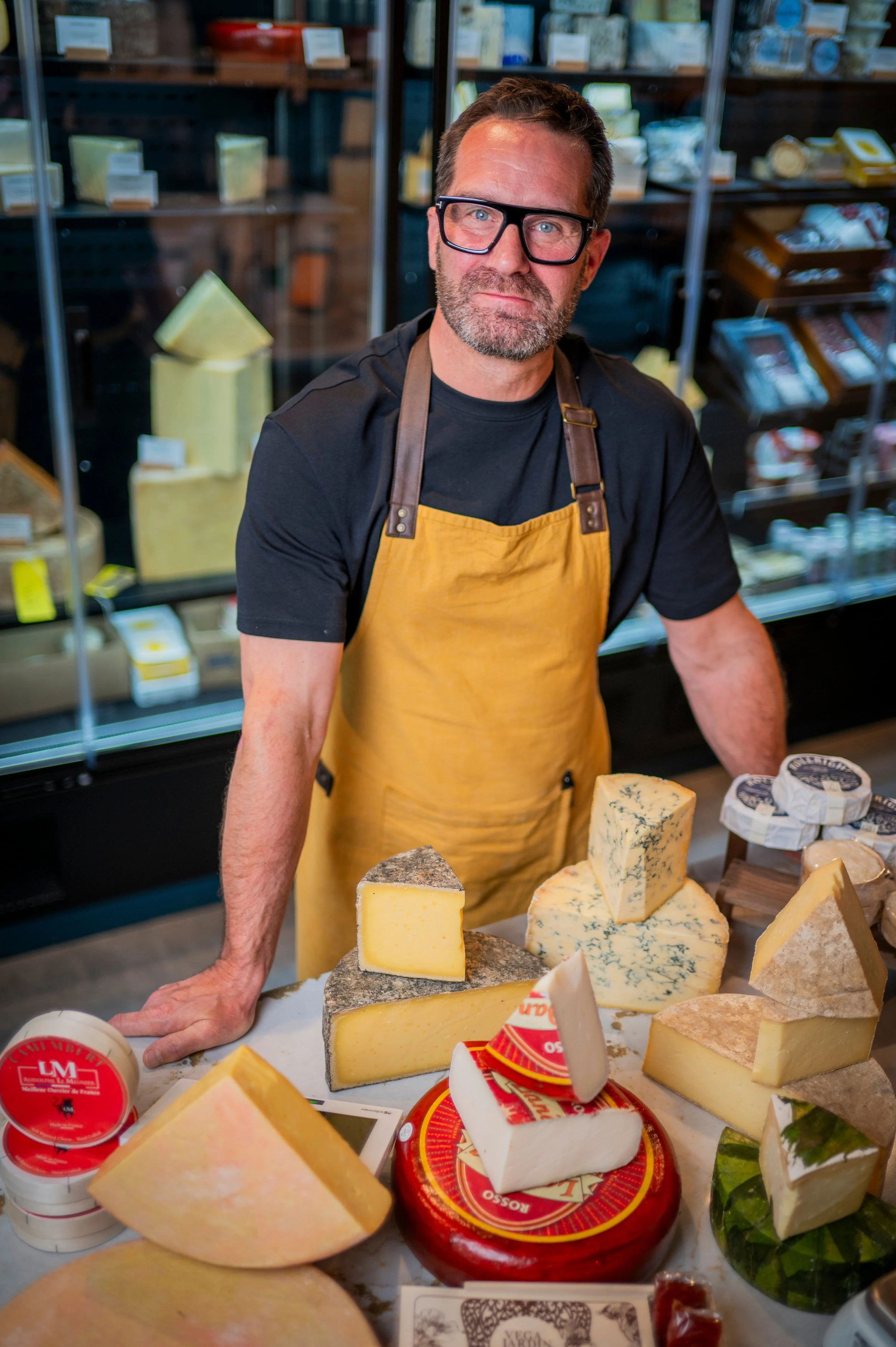 A man wearing glasses and a yellow apron behind a table with assorted cheeses and cheese containers, with shelves of packaged food in the background.