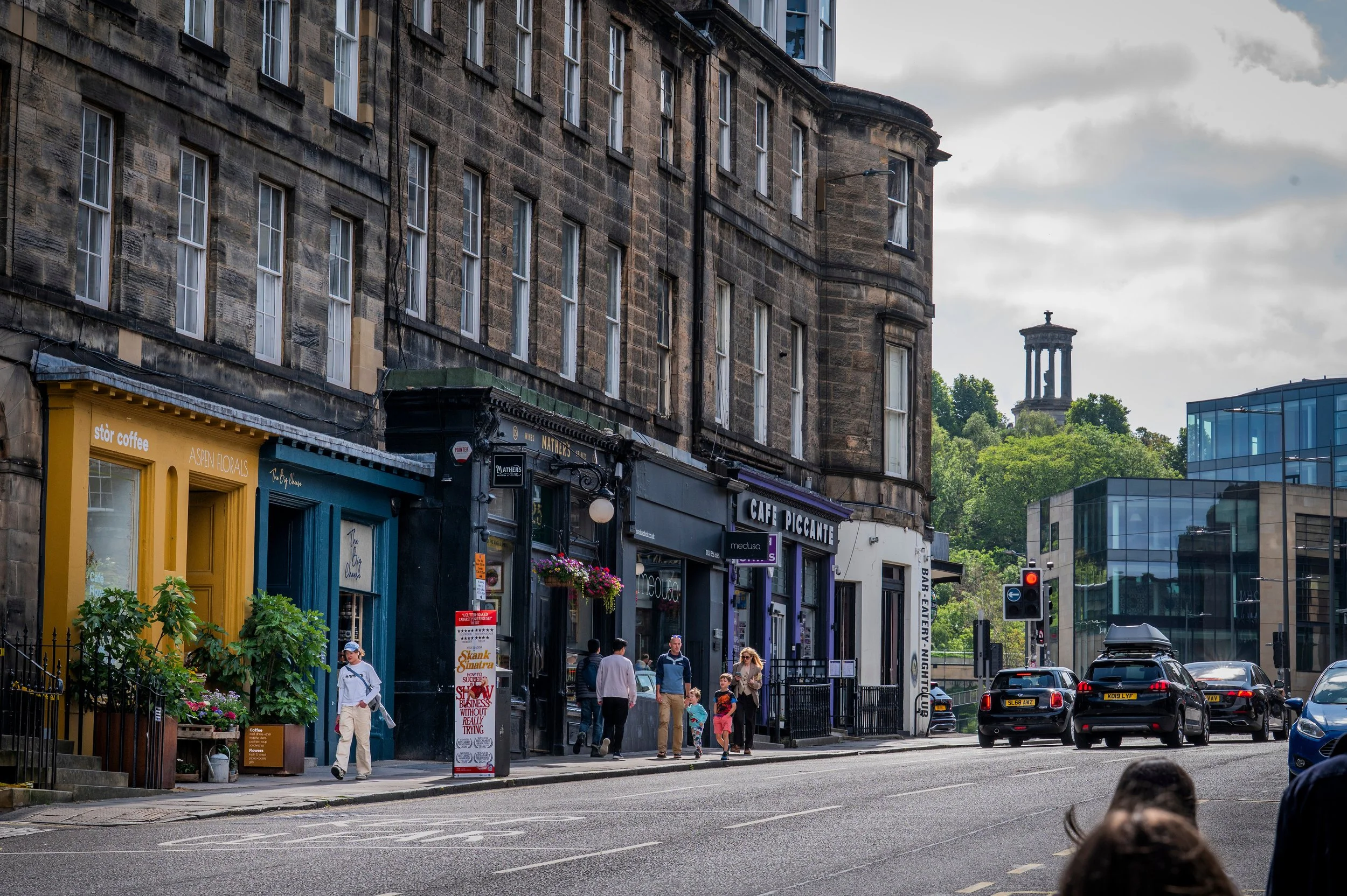 Street scene with people walking on the sidewalk, storefronts with colorful signs, parked cars on the street, and an old stone building with multiple windows in the background. A tower with classical architecture is visible atop a hill in the distance.