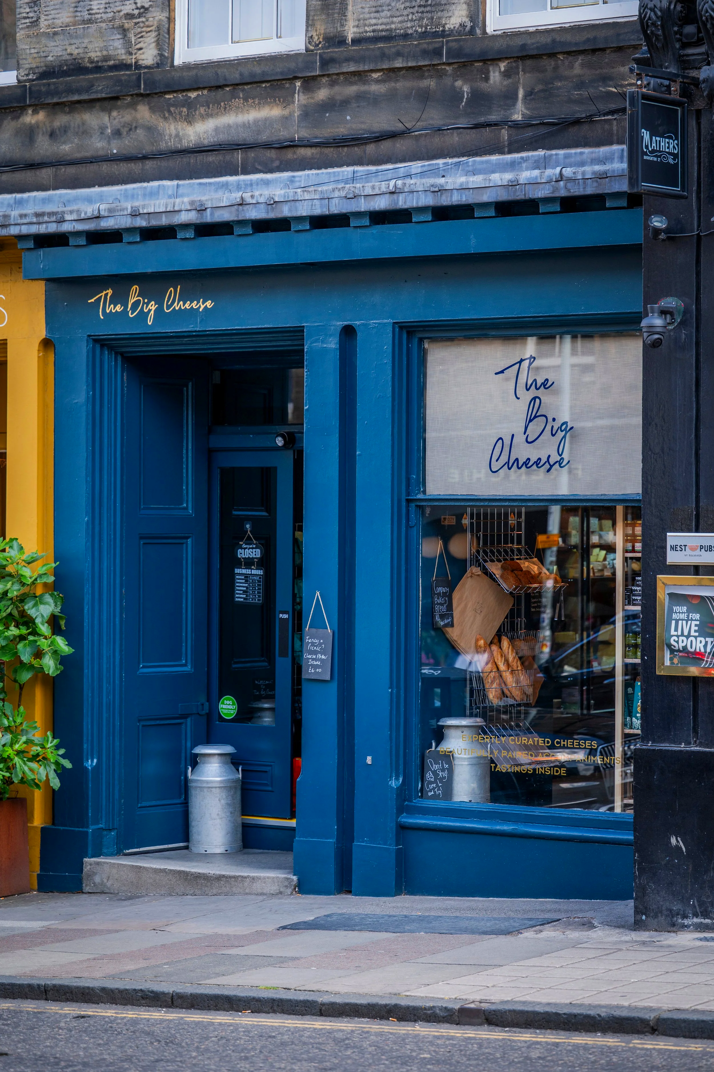 The storefront of a cheese shop called 'The Big Cheese' with a blue door and window. Inside, bread is displayed. A sign indicates it is closed, and another sign advertises curated cheeses with tasting inside. There is also a metal milk can on the sidewalk in front of the shop.
