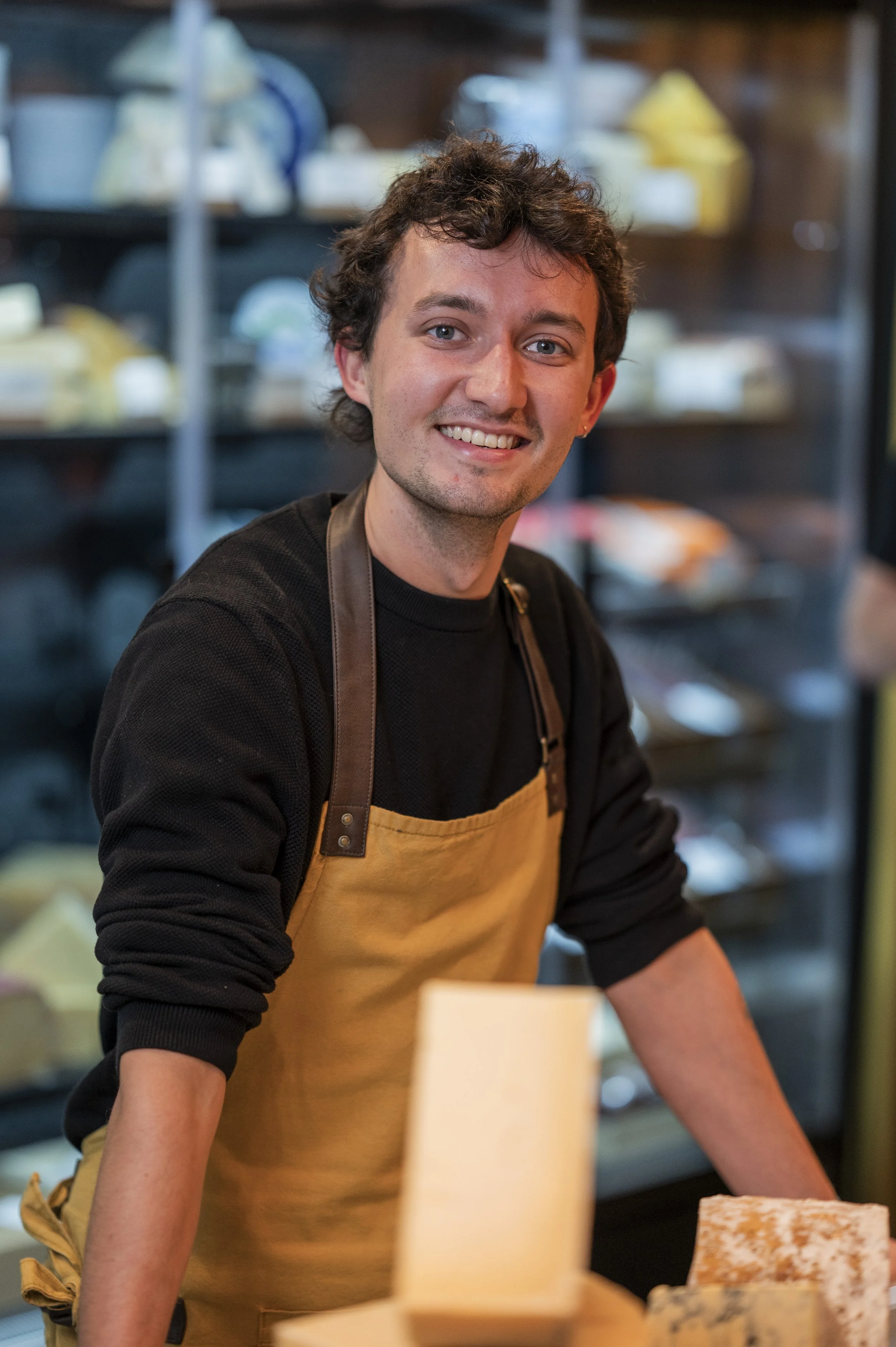 A young man with short curly hair and blue eyes smiling, wearing a black shirt and a tan apron with brown straps, standing in a cheese shop or deli with shelves of cheese and other food items in the background.