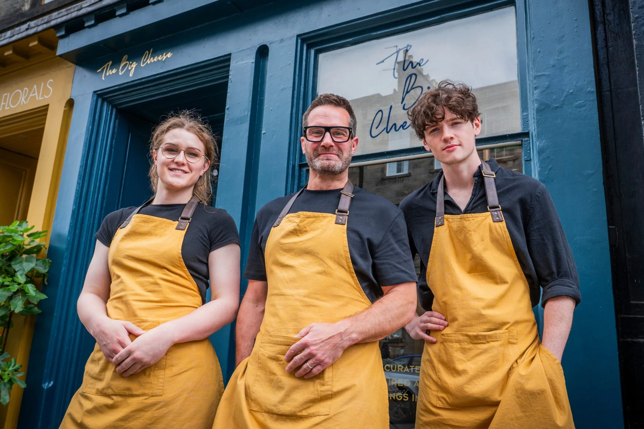 Three people standing outside a shop called 'The Big Cheese,' wearing black shirts and yellow aprons. The shop has a blue facade with a window and a sign.
