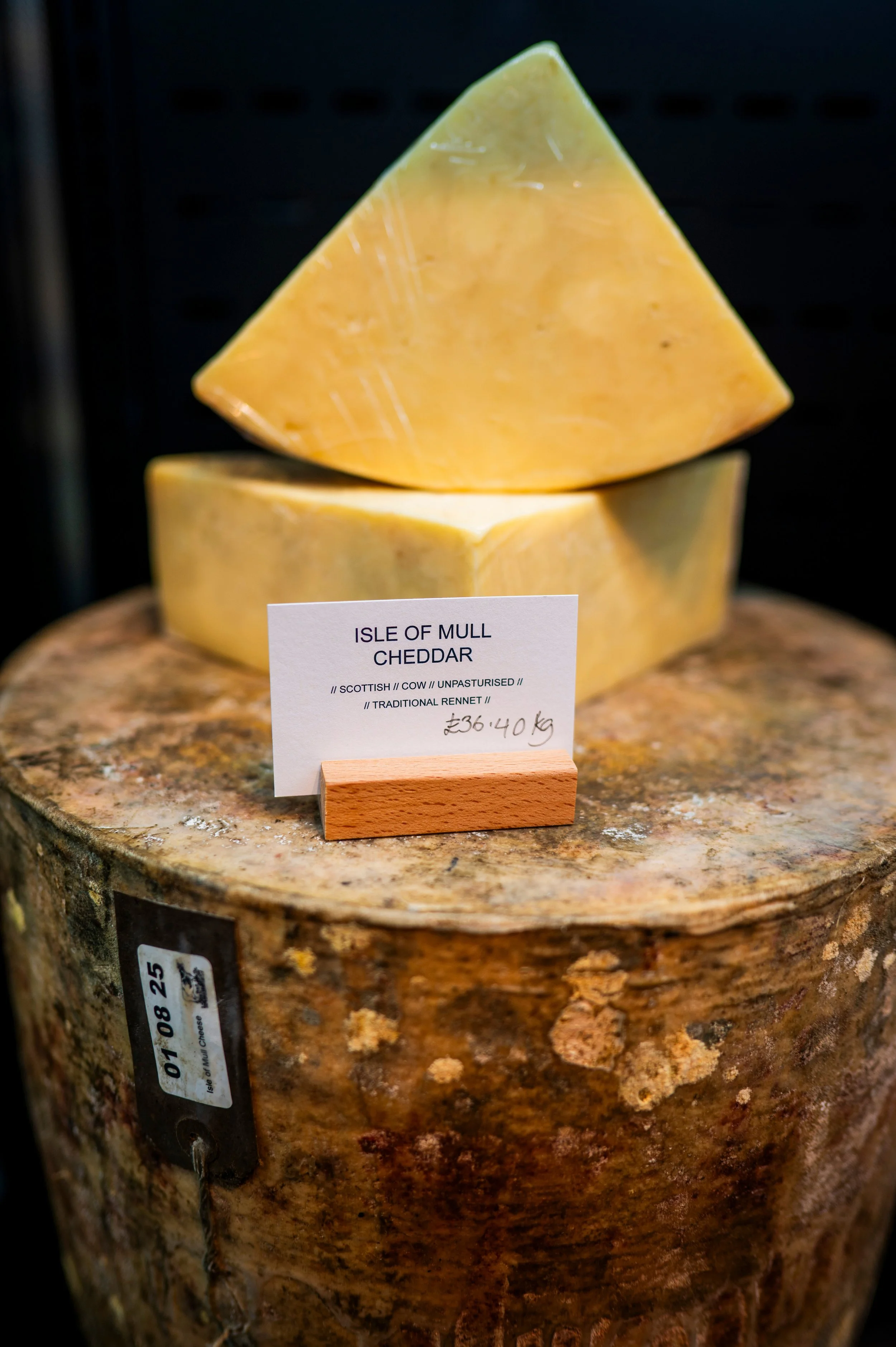 A block of Isle of Mull Cheddar cheese on display with a label in front of it. The cheese is yellow with a natural rind and is placed on a wooden surface.