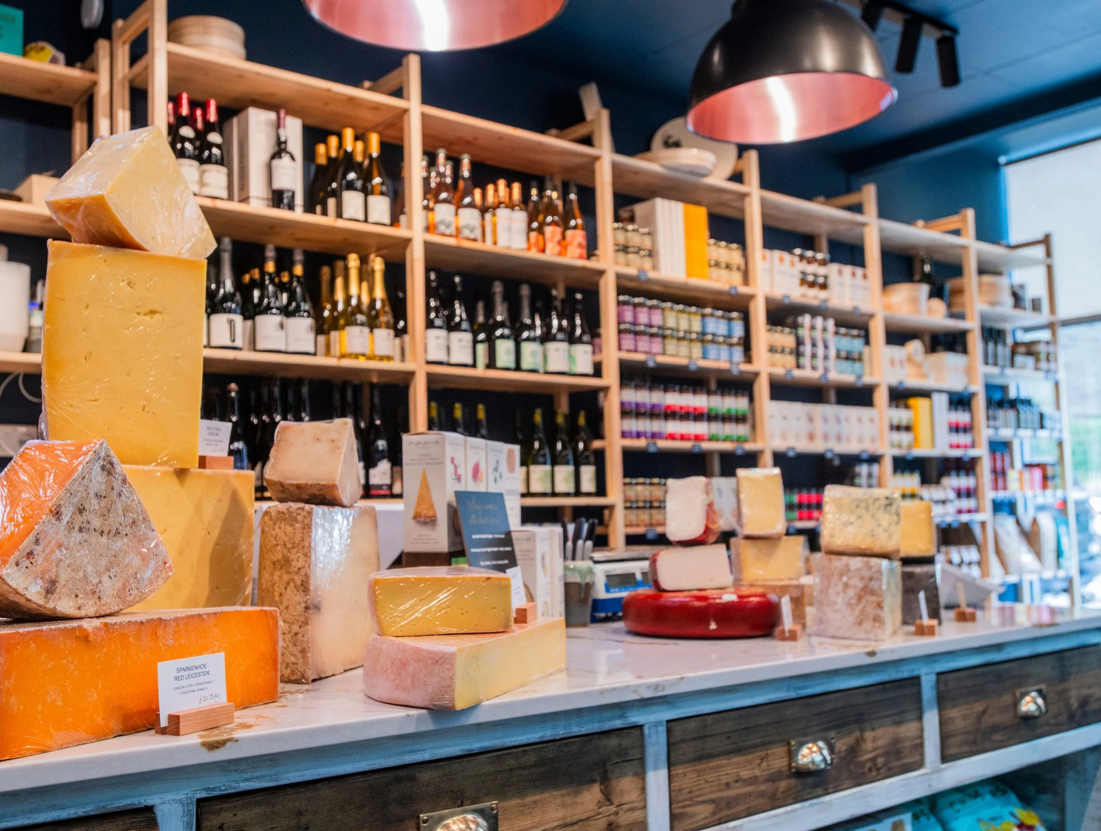 Display of various cheeses on a wooden counter in a store, with a shelf of wine bottles in the background.