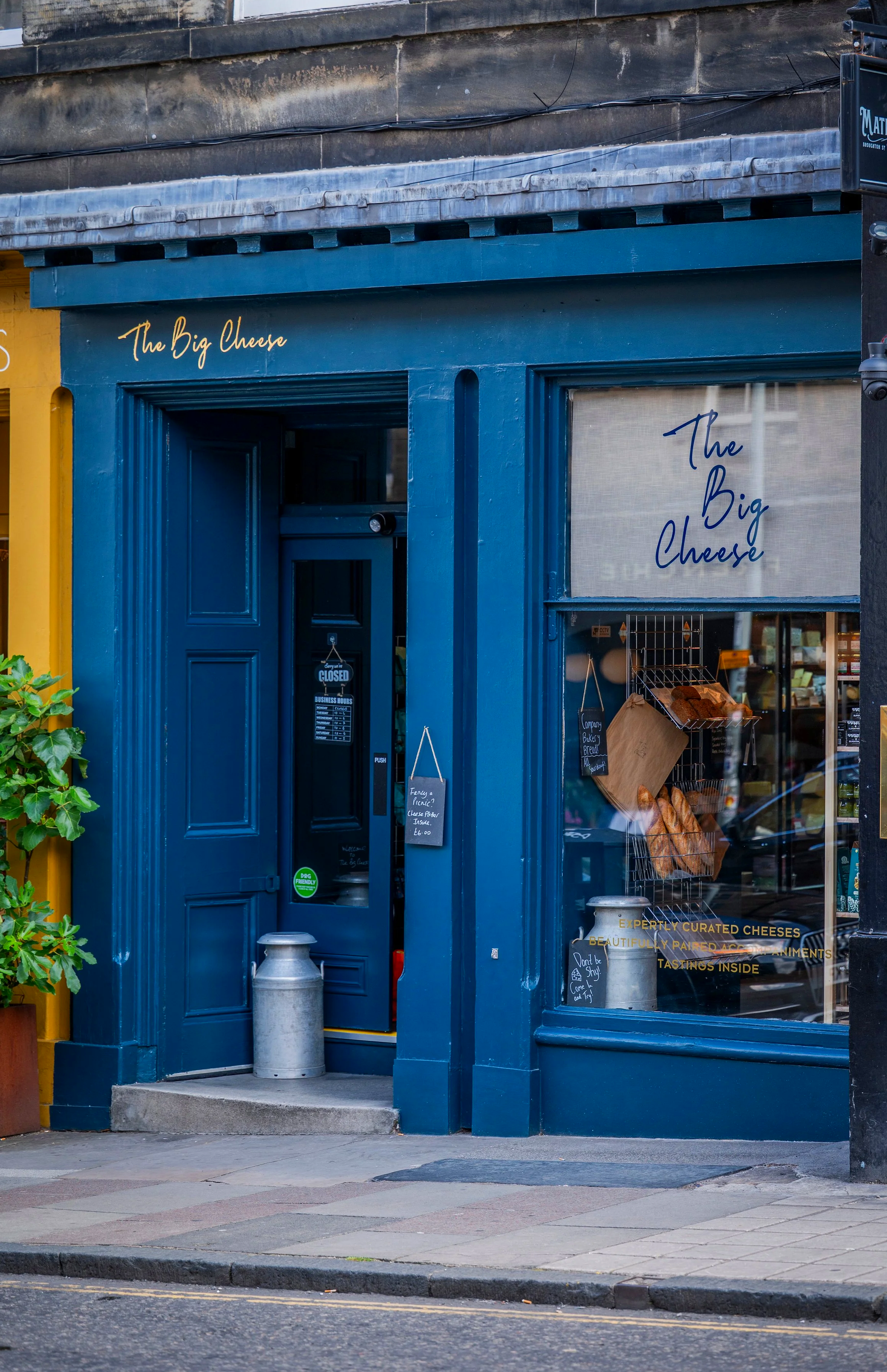 The exterior of a cheese shop with blue painted wood framing around the door and window. The shop sign reads "The Big Cheese" in cursive. Inside the window, Display shelves with cheeses and bakery items are visible, along with posters and signs promoting products and tasting events. Two metal bollards are outside the entrance, and a chalkboard sign is placed on the sidewalk.