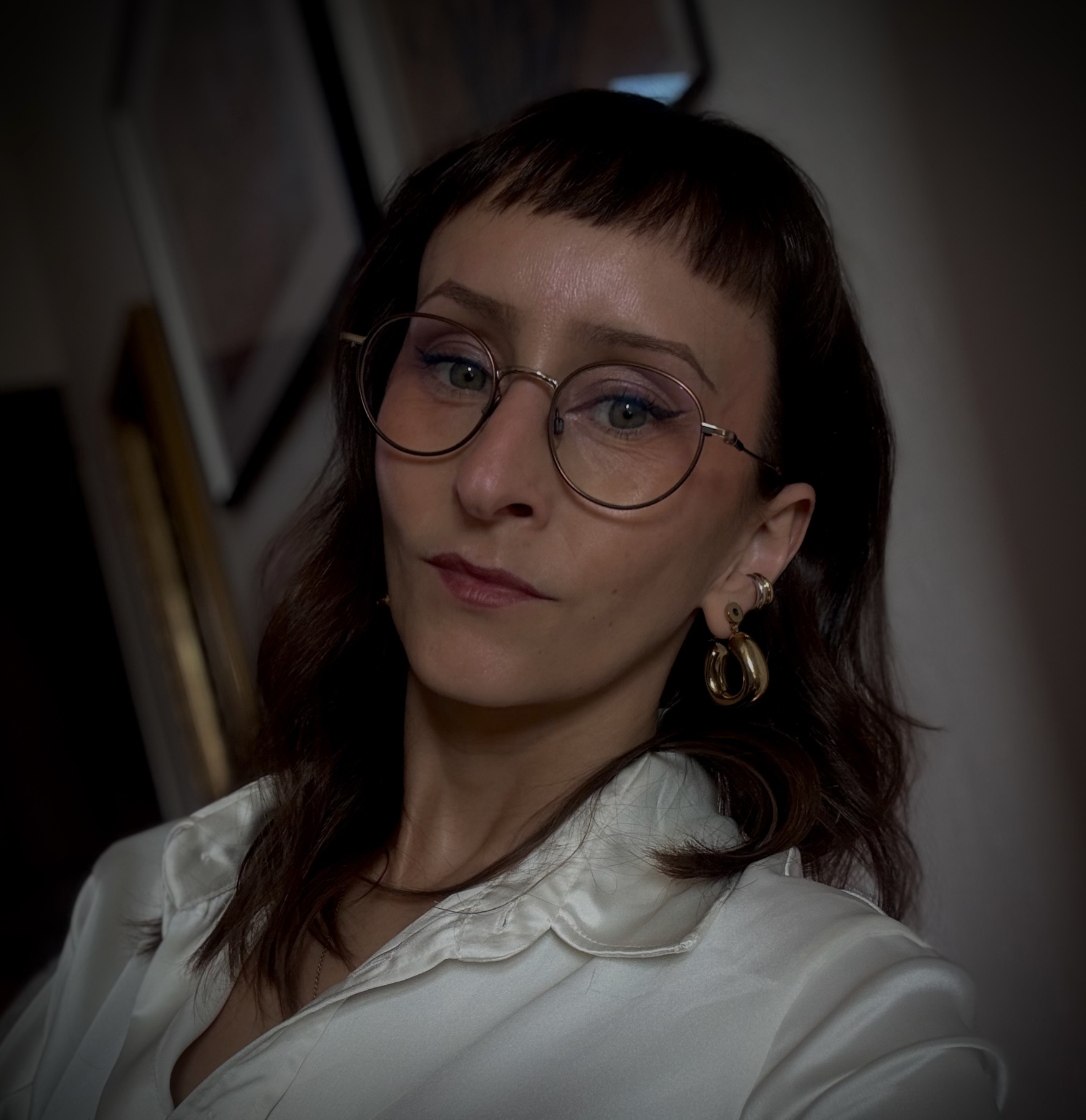 Une femme portant des lunettes rondes, des boucles d'oreilles dorées et une chemise blanche, regardant vers la droite, avec un fond sombre et flou.