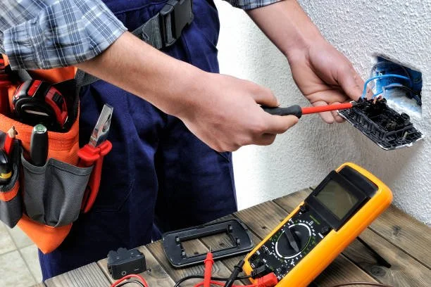 A person using a screwdriver to work on an electrical outlet with a multimeter on the table nearby.