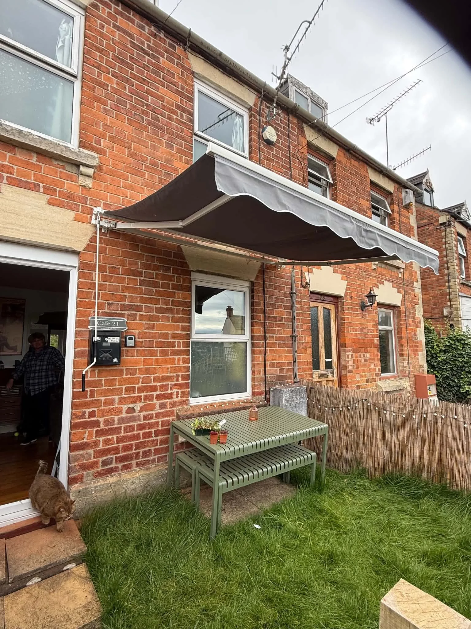The back patio of a red brick house with a black retractable awning, a green outdoor table, and a small grassy yard with a dog and a person visible through the open door.