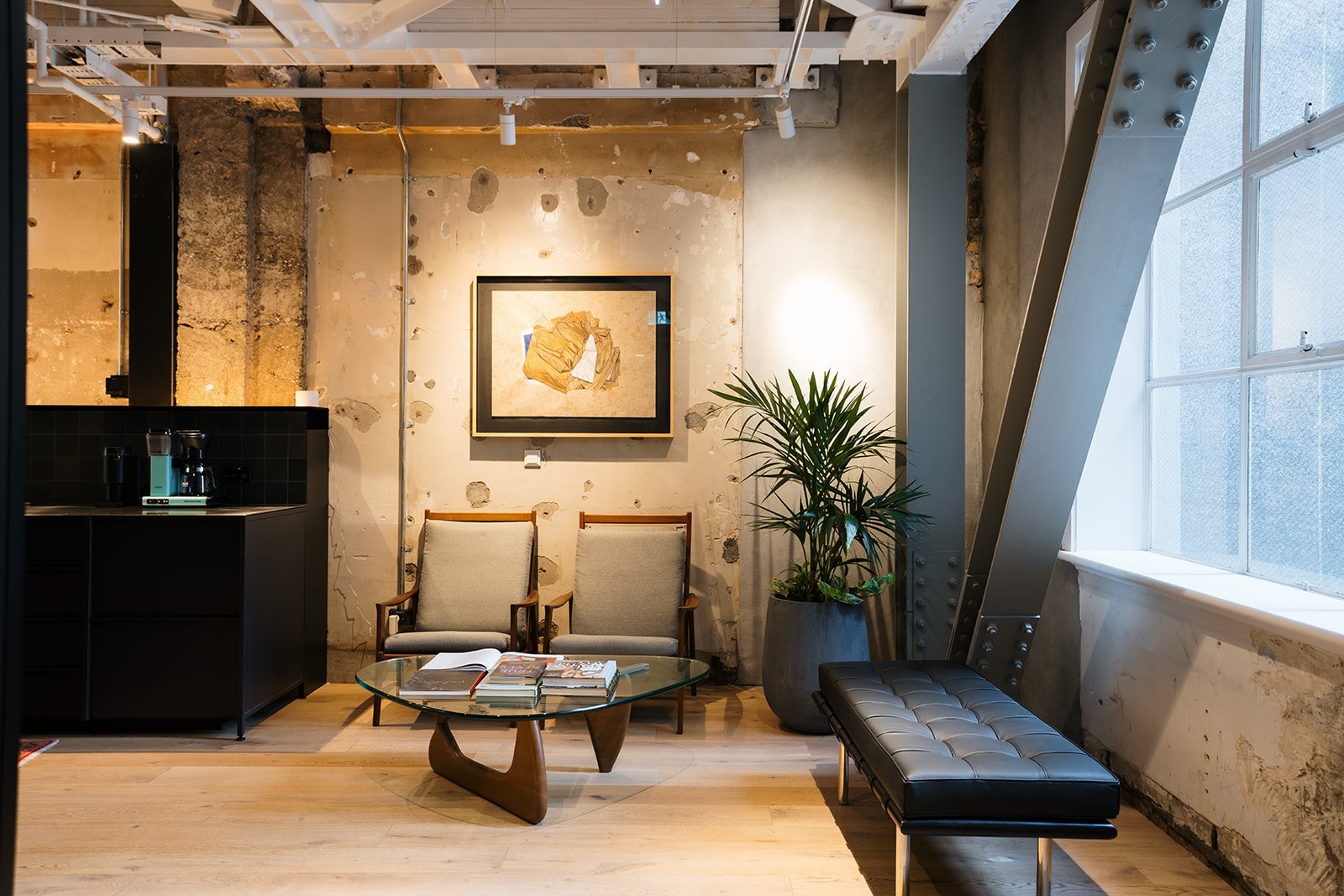 Interior of a modern loft-style room featuring two beige armchairs, a round glass coffee table with books, a potted plant, a black bench, and a wall with artistic decor, illuminated by natural light from large windows.