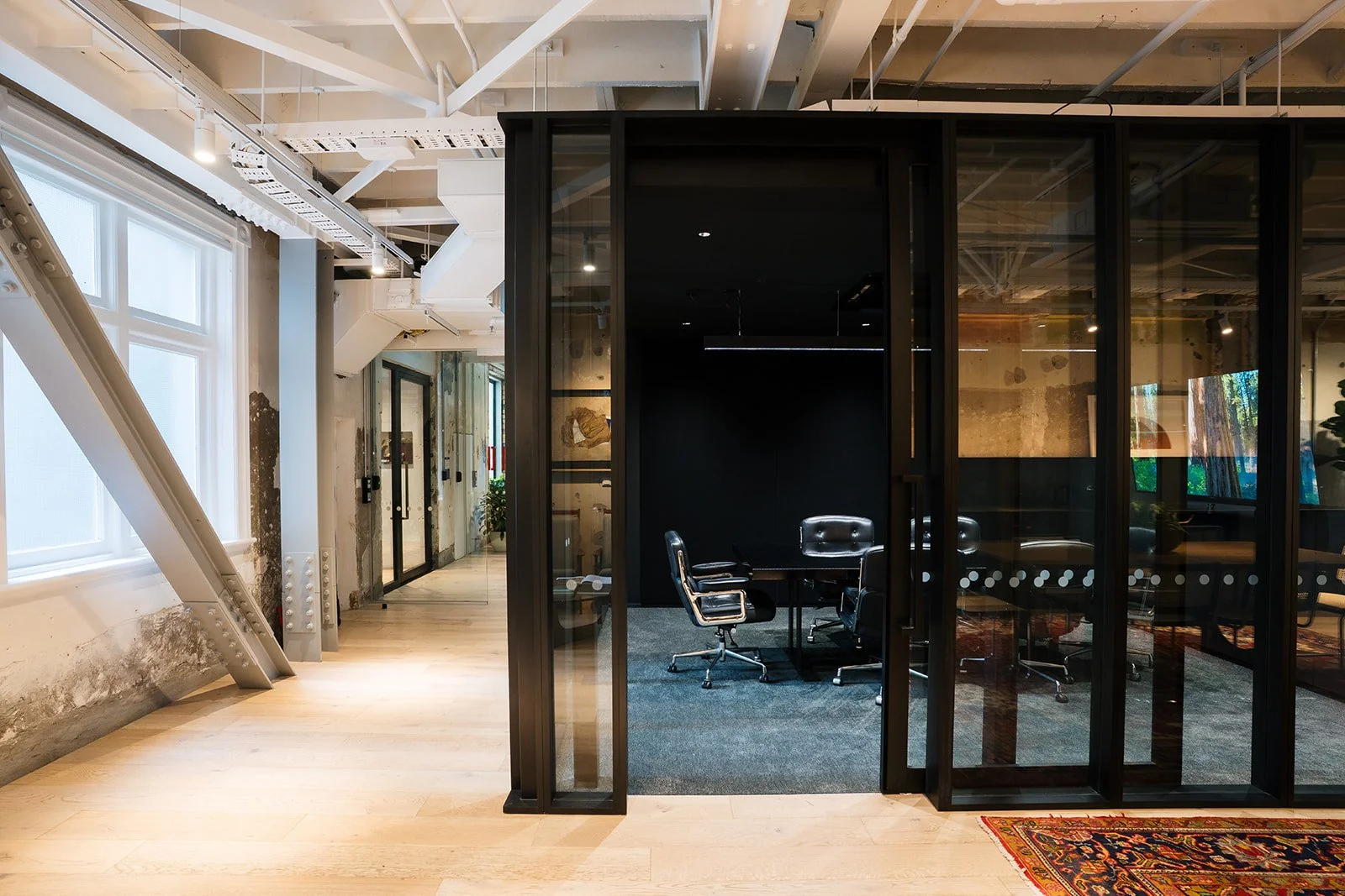 Modern office space with large windows, exposed ceiling pipes, and a glass-walled meeting room with black framing and black office chairs around a table.