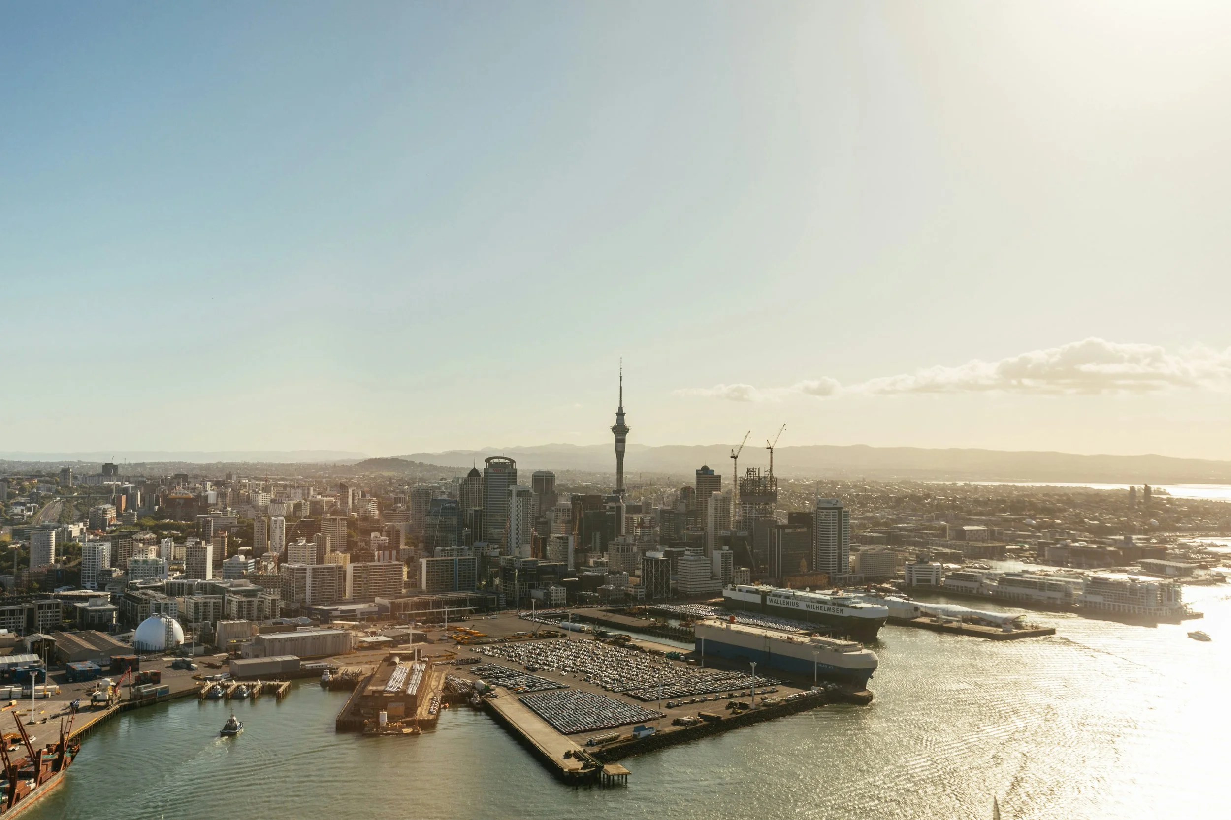 A city skyline with a tall tower in the center, several construction cranes, and a waterfront area with boats and a parking lot. The sky is mostly clear with some clouds.