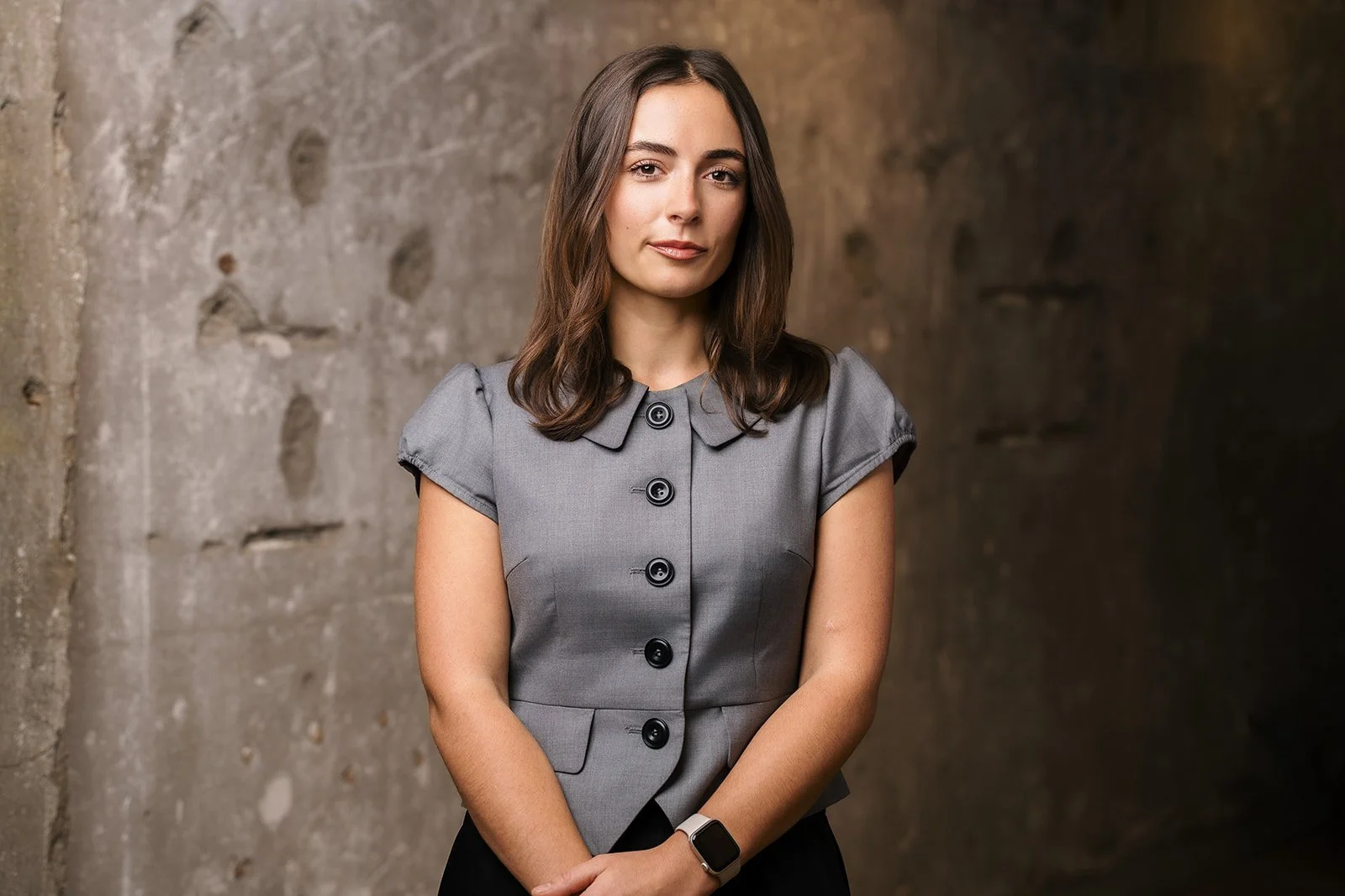 A woman with shoulder-length brown hair wearing a gray short-sleeved blazer with black buttons, standing against a textured concrete wall.