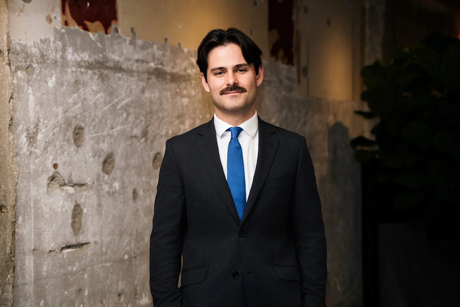 A man in a black suit, white shirt, and blue tie standing in front of a worn brick wall and dark foliage, smiling at the camera.