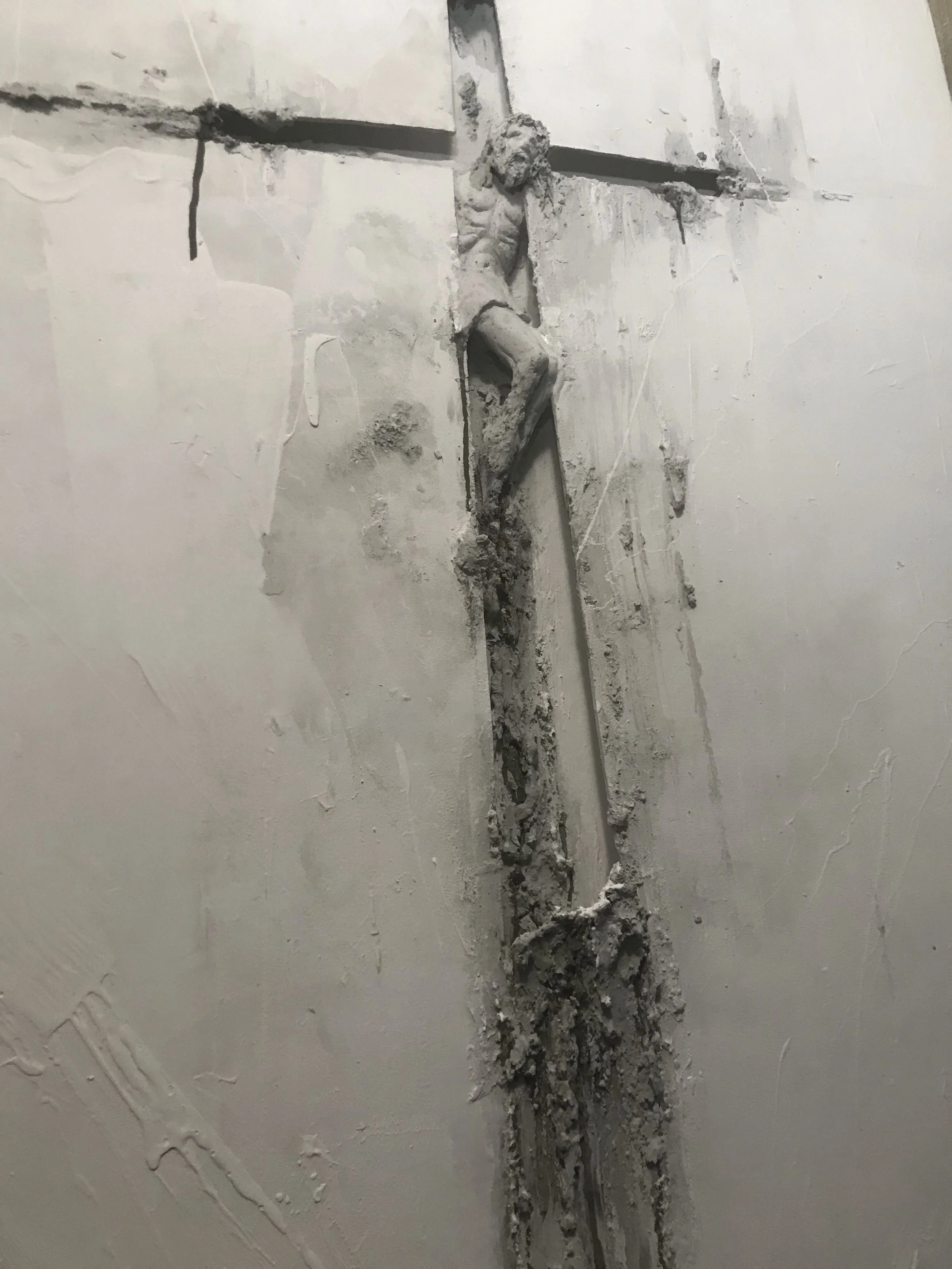 Statua di Gesù crocifisso in corso di restauro o deterioramento, con parti di cemento e malta visibili e alcune aree di esposizione del supporto originale.
