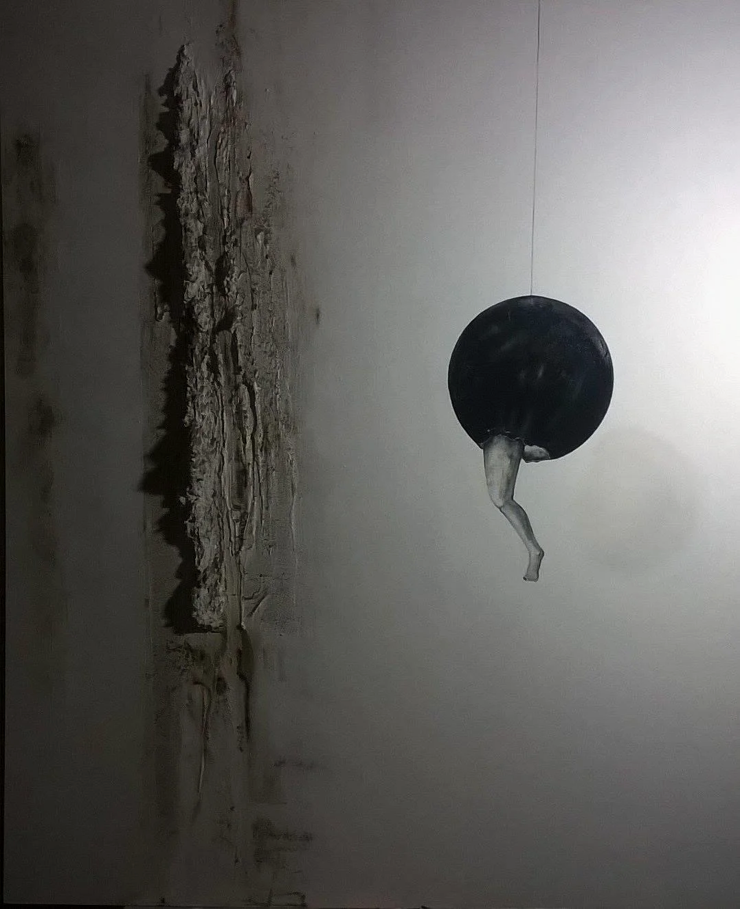 Installazione artistica di una figura umana che spunta da una sfera nera sospesa, con parete rotta e luci soffuse.