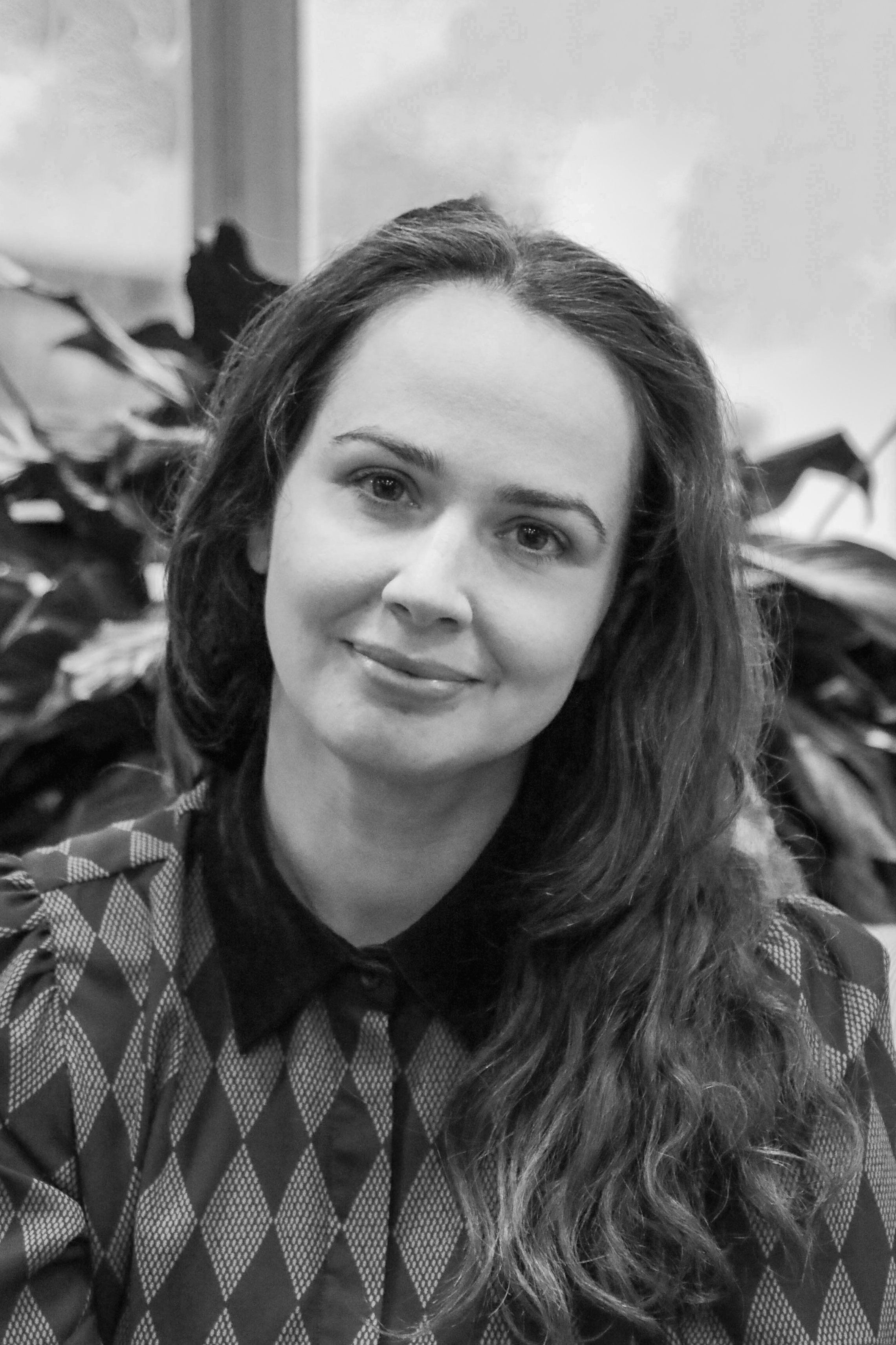 Black and white photo of a woman with long hair, wearing a patterned sweater, smiling softly at the camera, with plants in the background.