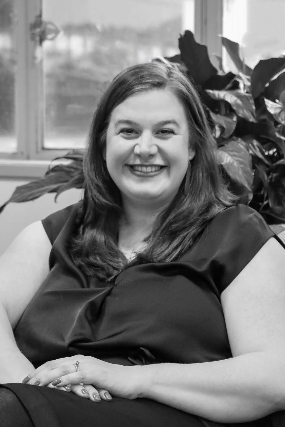 A woman with long hair smiling, sitting in front of a window and a large leafy plant, in black and white.