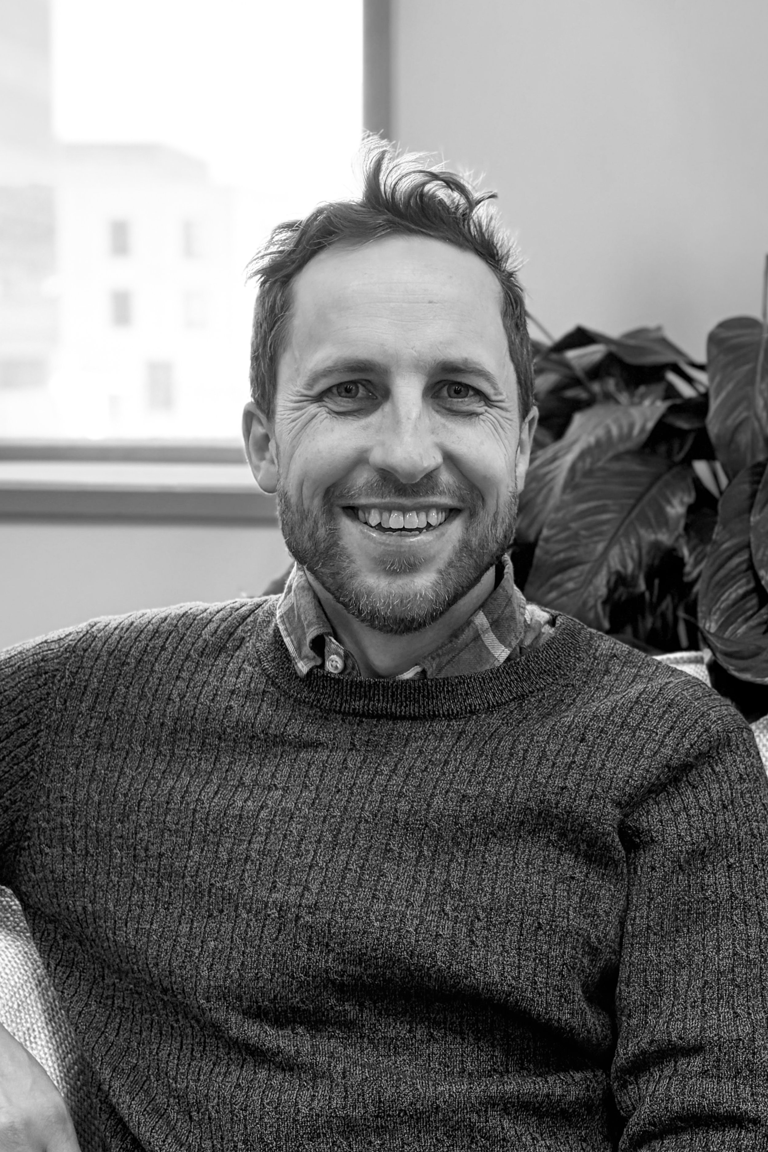Close-up black and white photograph of a smiling man with short, tousled hair and a beard, wearing a sweater and collared shirt, indoors with a window and plant in the background.